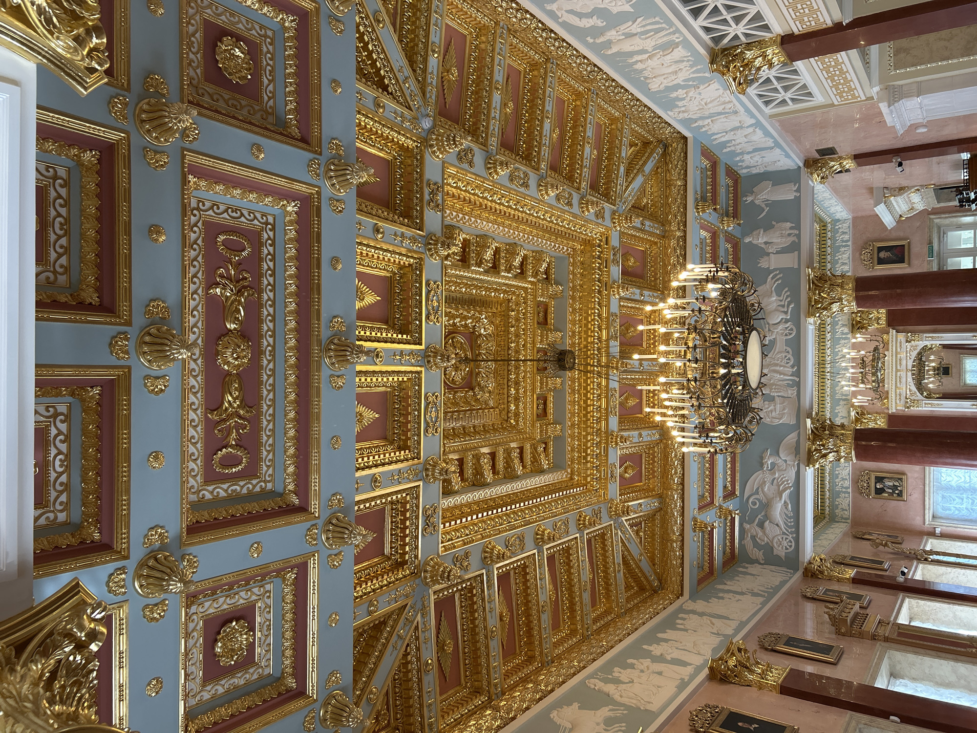 The image shows an opulent interior ceiling decorated with intricate gold ornamentation and a light blue and pink color scheme. The ceiling is divided into square and rectangular coffers, each featuring detailed gold relief work. A large, multi-tiered chandelier with numerous lit candles hangs from the center of the ceiling. The walls are a pale pink and feature classical friezes with figures in relief. Large, dark red columns with gold capitals support the upper structures. Several framed portraits are visible on the walls, and windows with intricate white frames are present. The overall impression is of a grand and historically significant room, likely a palace or a museum. The specific location in Добасненскі сельскі Савет, Belarus, is not evident from the visual information provided in the image.