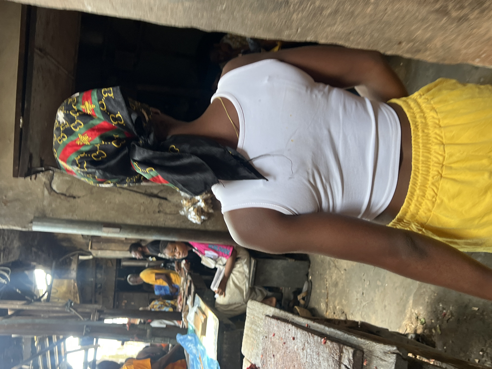 The image shows a young Black female from behind, wearing a white tank top and bright yellow velour shorts. She has a headscarf with a black, green, red, and gold pattern, tied with a knot at the back. Her arm is visible, showing darker skin on the elbow. The background suggests an outdoor market setting in Nkpor, Nigeria. To the left, a wooden table with what appears to be raw meat and a blue plastic sheet is visible. Further back, several people are seen at stalls, including a woman in a pink top sitting and reading a book, and men in yellow shirts. The environment is rustic with rough concrete walls and wooden beams overhead, indicating a possibly covered but open-air market. The lighting is natural and diffused.