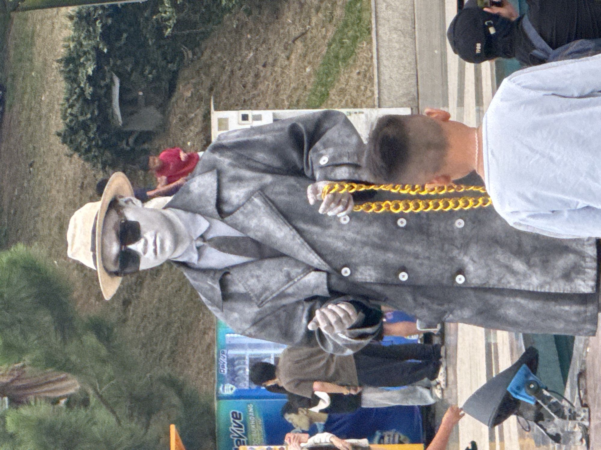 In Da Lat, Vietnam, a man painted silver in a trench coat and fedora stands on a paved area. He wears sunglasses and holds a large gold chain. Behind him, a young boy in a light blue shirt looks away. To the right, another person wears a black cap and backpack, holding a camera. In the background, people in casual clothing mill about near a vending machine and green foliage.