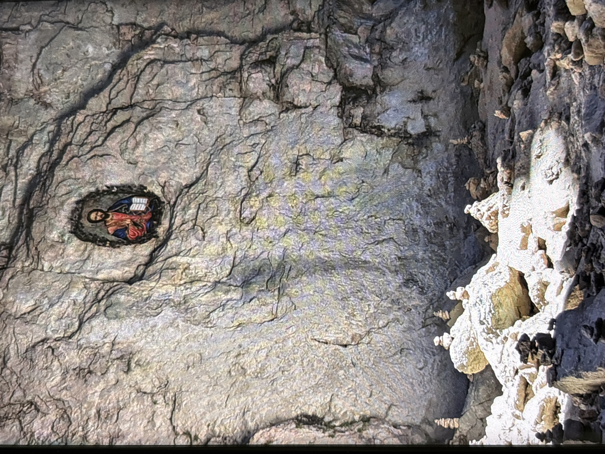 The image presents a vertically oriented, close-up view of a rugged, rock-hewn interior, characterized by deep textures and irregular surfaces. It appears to have been rotated 90 degrees clockwise from its original capture.
On the left side of the frame, a religious icon is prominently displayed within a dark, roughly circular recess in the rock face. The icon features a bearded figure, likely Jesus Christ or a saint, adorned with a halo and robes of red and blue. The figure holds an open book, its pages showing indistinct markings, and their gaze is directed downwards (as viewed in the current vertical orientation). The surrounding rock is a mottled gray with discernible veins and darker crevices.
The central and upper portions of the image consist of extensive rock surfaces, marked by intricate patterns of dark fissures and lighter, occasionally iridescent patches, indicating varied mineral deposits or reflective qualities.
The right side of the frame showcases a complex array of lighter-colored, irregular formations, reminiscent of stalactites or other cave concretions. These protrusions, ranging from off-white to yellowish hues, exhibit stark contrasts of light and shadow, emphasizing their three-dimensional and often crystalline or encrusted textures.
The scene is static, depicting an artistic or religious installation within a natural or carved rock setting. Directed lighting creates dramatic highlights and shadows across the surfaces. No distinct text is legible within the image, beyond the vague markings on the figure's book. The provided context states the location as Wolverhampton, United Kingdom, suggesting this is a feature within that area.
