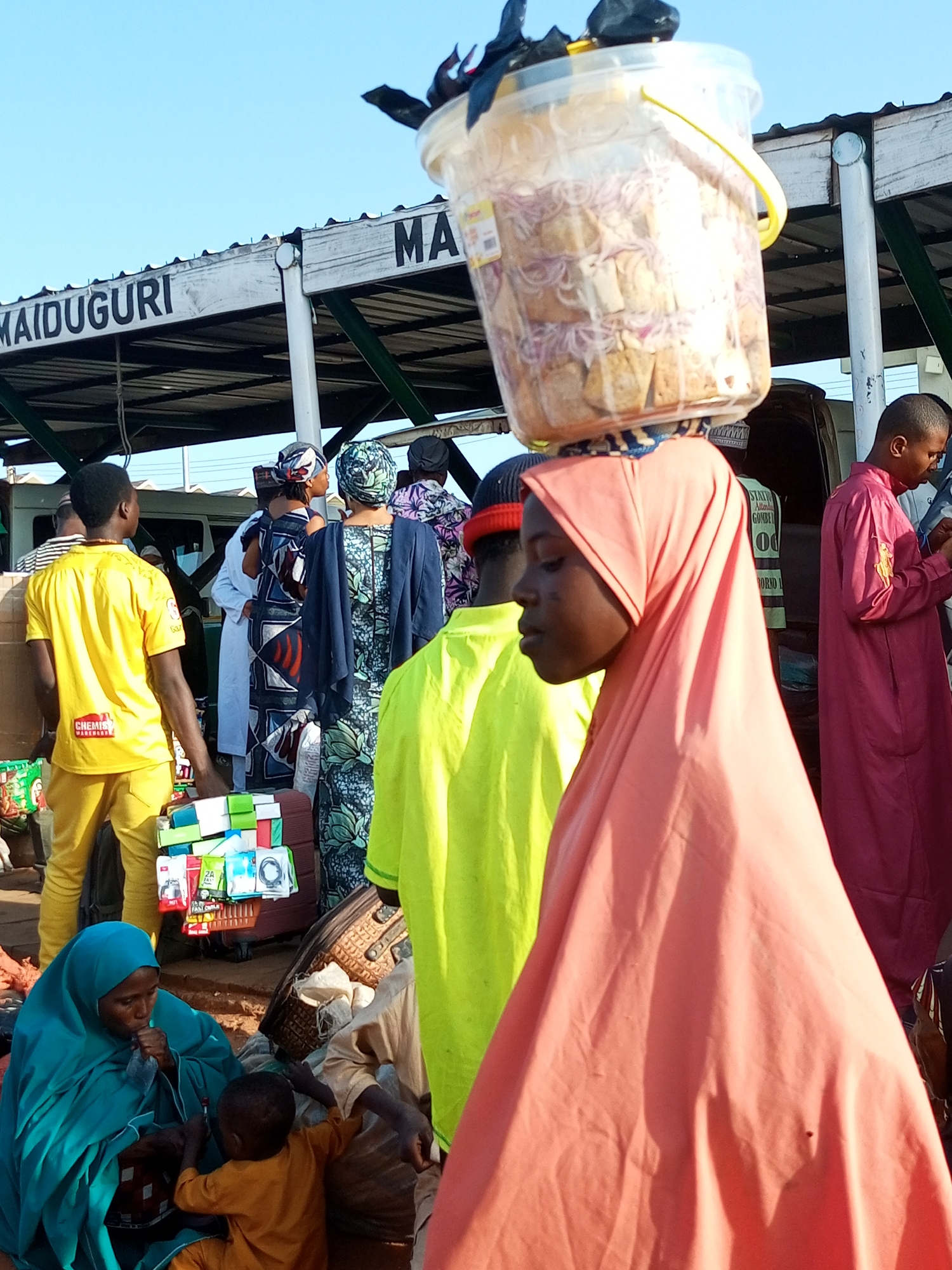 This is a street-level photograph taken in Maiduguri, Gombe, Nigeria, likely at a transportation hub or market. The scene is bustling with activity under a clear blue sky and a covered walkway with signage that reads "MAIDUGURI".
In the foreground, a young girl in a bright coral-colored hijab is the closest subject, her face in profile. She carries a large, transparent plastic bucket filled with food items, possibly bread and onions, balanced on her head. The bucket has a yellow handle and some black debris on top.
To her left, a woman in a teal hijab is seated, holding a young child wearing an orange outfit. The woman appears to be eating or drinking from a plastic package. The child is reaching towards her.
Further back, several other individuals are visible. A man in a bright yellow t-shirt with "CHEMIST WAREHOUSE" printed on it stands facing away from the camera, carrying what appear to be boxes or packages. Adjacent to him, a stack of various small packaged goods, possibly phone accessories, is displayed on a stand.
Other people are seen in traditional attire, including a woman in a patterned blue and white dress and another in a dark blue cloak. A man in a pink traditional robe stands on the right side of the frame, looking down at his phone.
The environment suggests a busy public space with a covered waiting area or stalls. The ground is uneven and appears to be dirt or gravel. Bags and luggage are scattered around, indicating travel or trade.