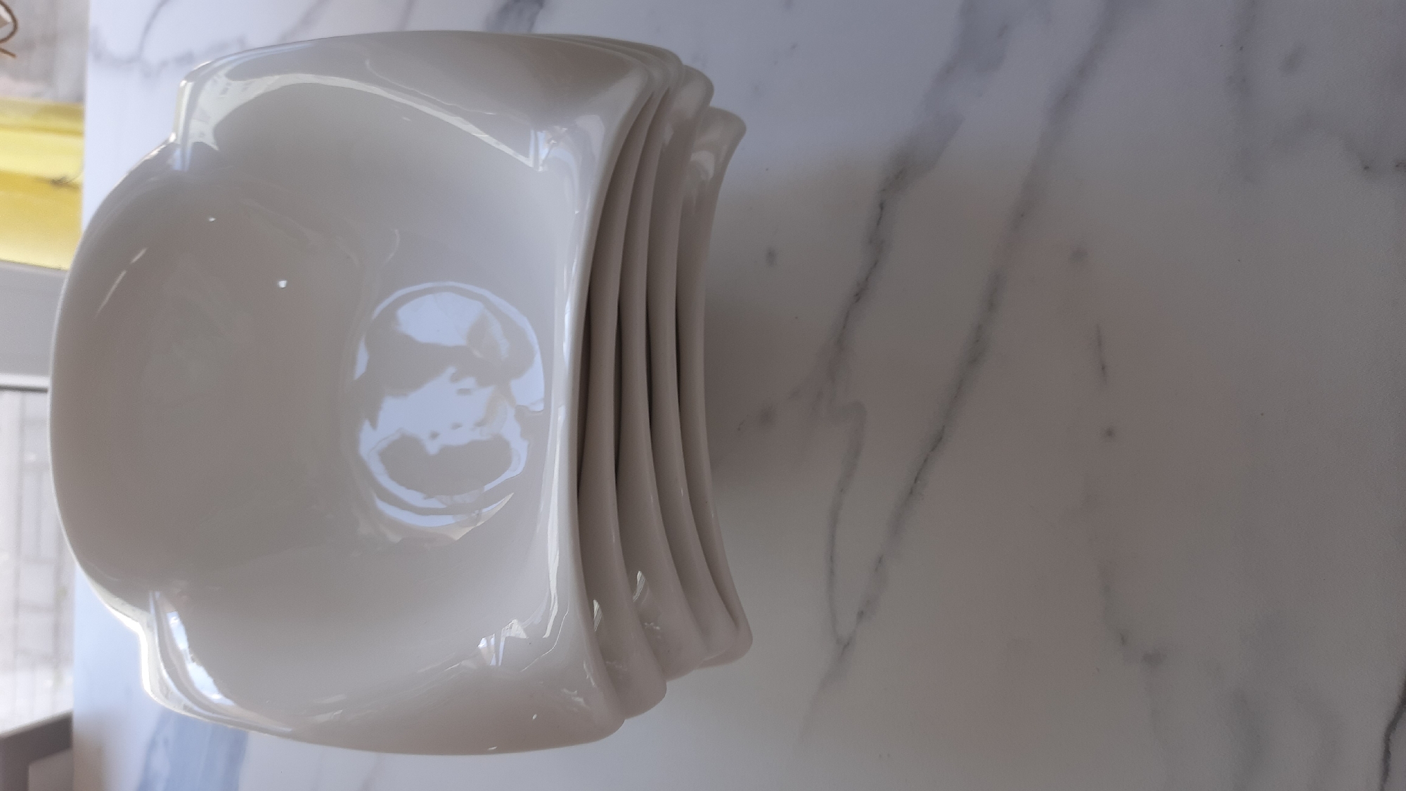 A stack of five white, glossy ceramic bowls is presented at an angle on a marble-patterned surface. The bowls have a unique, slightly wavy or sculpted edge, with each bowl nesting into the one below it. The lighting creates highlights and reflections on the smooth, curved surfaces of the bowls, emphasizing their shape and sheen. The marble surface has subtle grey veining, providing a neutral and elegant backdrop. The overall impression is one of a clean, modern aesthetic, likely within a domestic setting. The image does not depict any people, text, or specific indications of time of day or weather. The location context of Desnogorsk, Russia, does not provide any visible cues within the image itself.