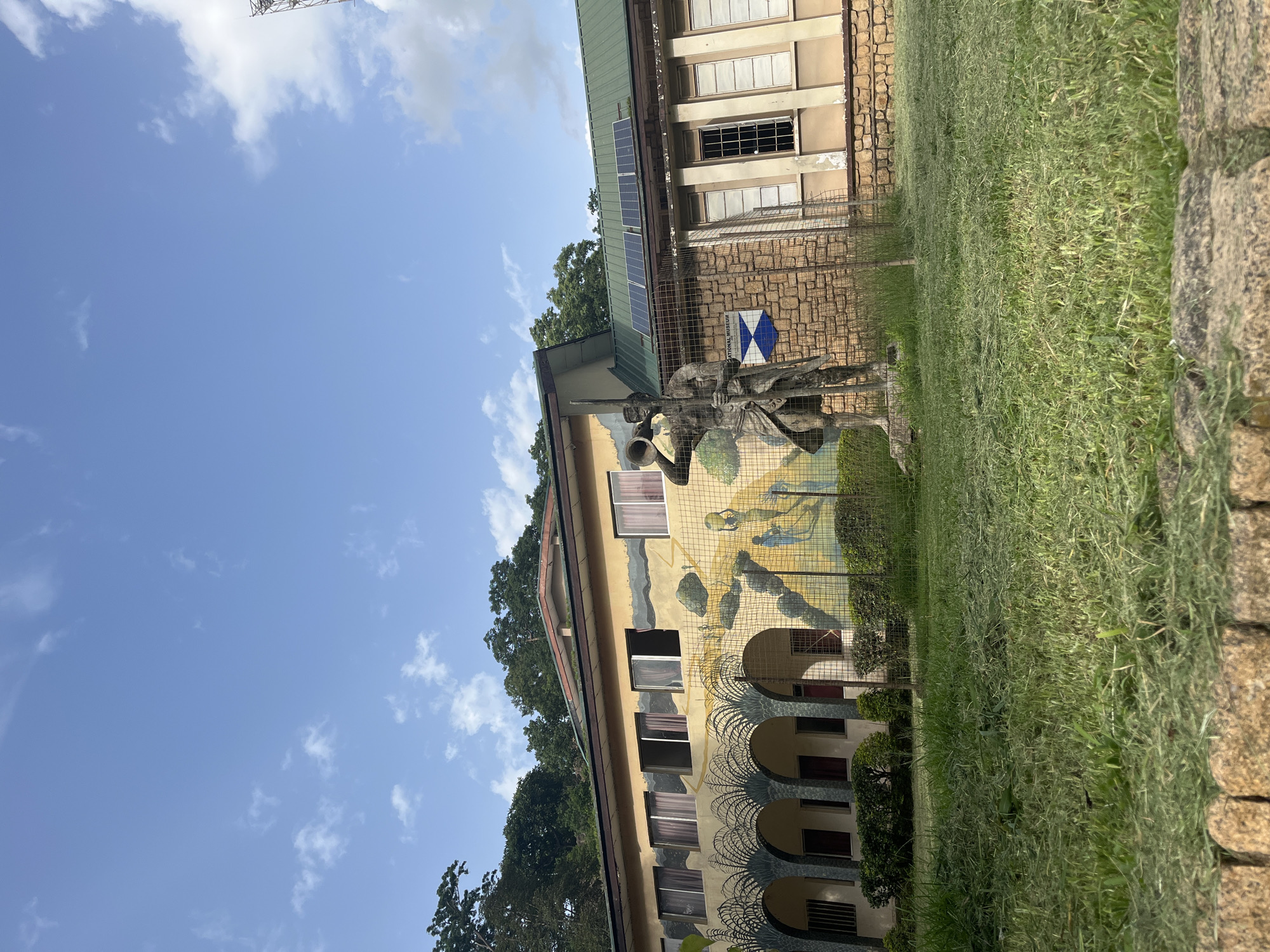 This image shows the exterior of the National Museum in Jos, Nigeria. The museum is a two-story building with a light yellow facade. The lower story features a mural depicting various scenes, including palm trees, people carrying water, and figures in a landscape. An arched colonnade runs along the front of this section. The upper story has several windows and is partially covered by a green roof. Solar panels are visible on the roof.
In front of the museum, on the lawn, stands a weathered statue of a figure holding a horn or trumpet and a spear. A wire fence encloses the statue and a portion of the museum's grounds. The foreground consists of a well-maintained grassy lawn with cut grass scattered on it. In the background, lush green trees and a bright blue sky with scattered clouds complete the scene.