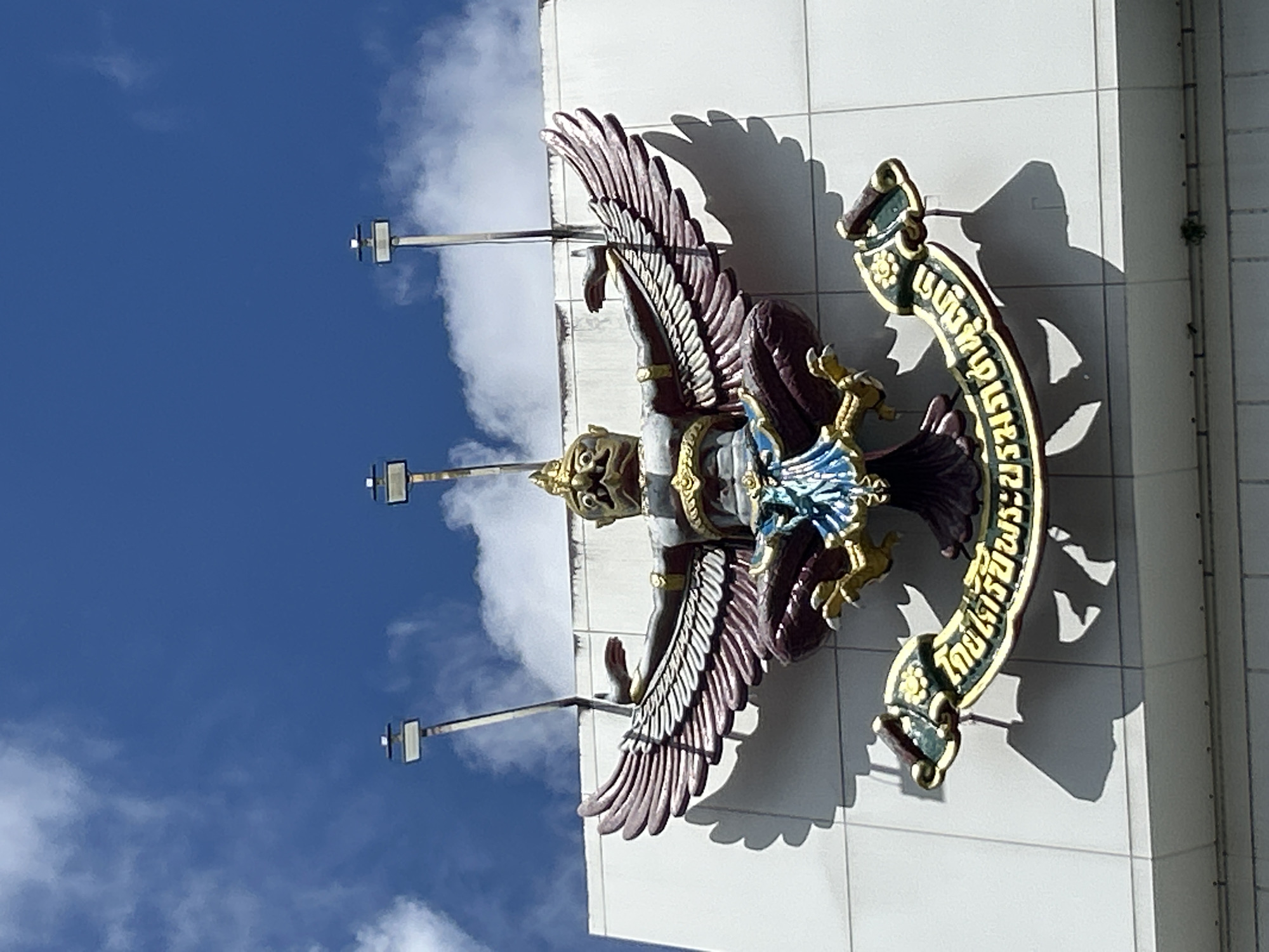 A statue of a Garuda, a mythical bird-like creature, is mounted on the facade of a white building. The Garuda is depicted with outstretched wings, a human-like torso, and an eagle's head. It is adorned with gold and blue accents. Below the Garuda, a scroll-like banner with Thai inscriptions is visible. Three modern spotlight fixtures are mounted above the statue. The background consists of a bright blue sky with scattered white clouds. The building appears to be located in Wichit, Thailand, as suggested by the style of the Garuda and the Thai inscription on the banner. The lighting indicates it is daytime.