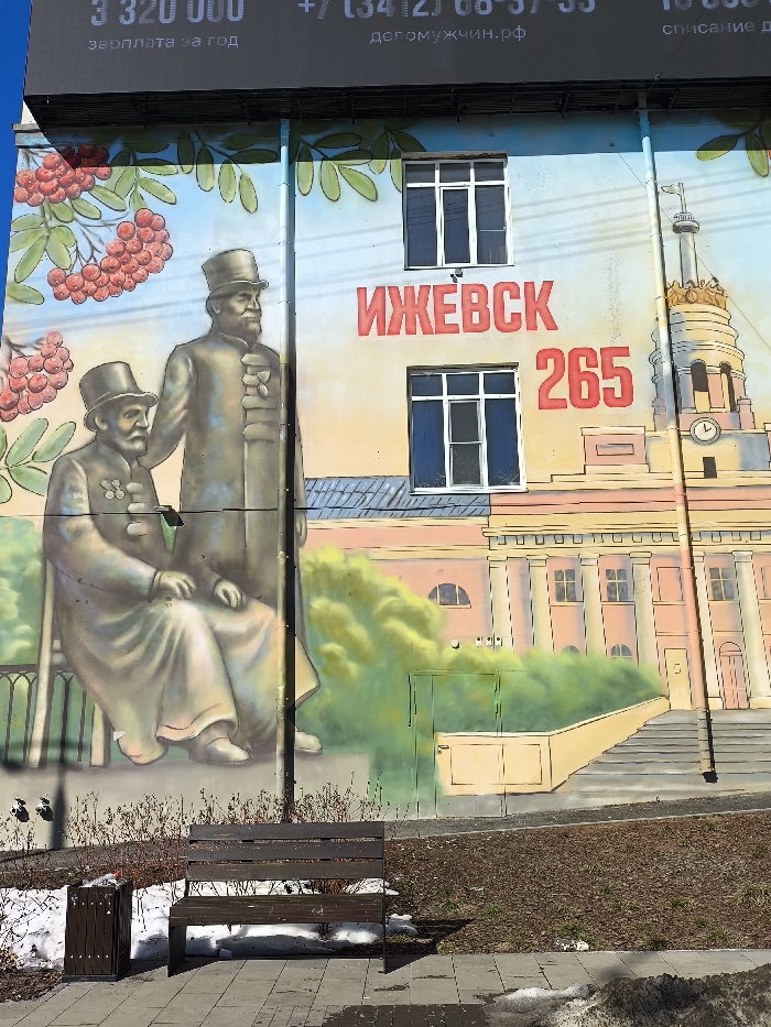 The image presents an outdoor urban scene dominated by a building wall covered in a large, colorful mural, beneath a black billboard. The mural, rendered in a stylized, illustrative manner, depicts several elements. On the left, clusters of red berries and green leaves are painted. Towards the center-left, two men in 19th-century attire, including top hats, beards, and coats with medals, are illustrated; one is seated on a bench, and the other stands behind him. Centrally, two white-framed windows are integrated into the mural. The mural prominently features the large red Cyrillic word "ИЖЕВСК" (Izhevsk) and the red numerals "265". To the right, a detailed depiction of a classical-style building with columns, a grand staircase, a large clock, and a distinctive spire is visible.
Above the mural, a black billboard displays white Cyrillic text including "3 320 000", "зарплата за год" (salary per year), a phone number "+7 (3412) 68-57-55", and "деломужчин.рф".
At ground level, a dark brown wooden slatted bench with metal legs rests on a paved sidewalk. To its left, a matching dark brown wooden trash receptacle is situated. The surrounding ground cover consists of dry, dark earth and sparse, bare vegetation, interspersed with patches of melting, dirty snow, indicating cold weather conditions. A dark utility pole extends vertically from the ground, obscuring a small portion of the mural on the right side. The scene is illuminated by bright natural daylight. The mural itself depicts "Izhevsk," which is distinct from the user's specified city of Березник, Russia.