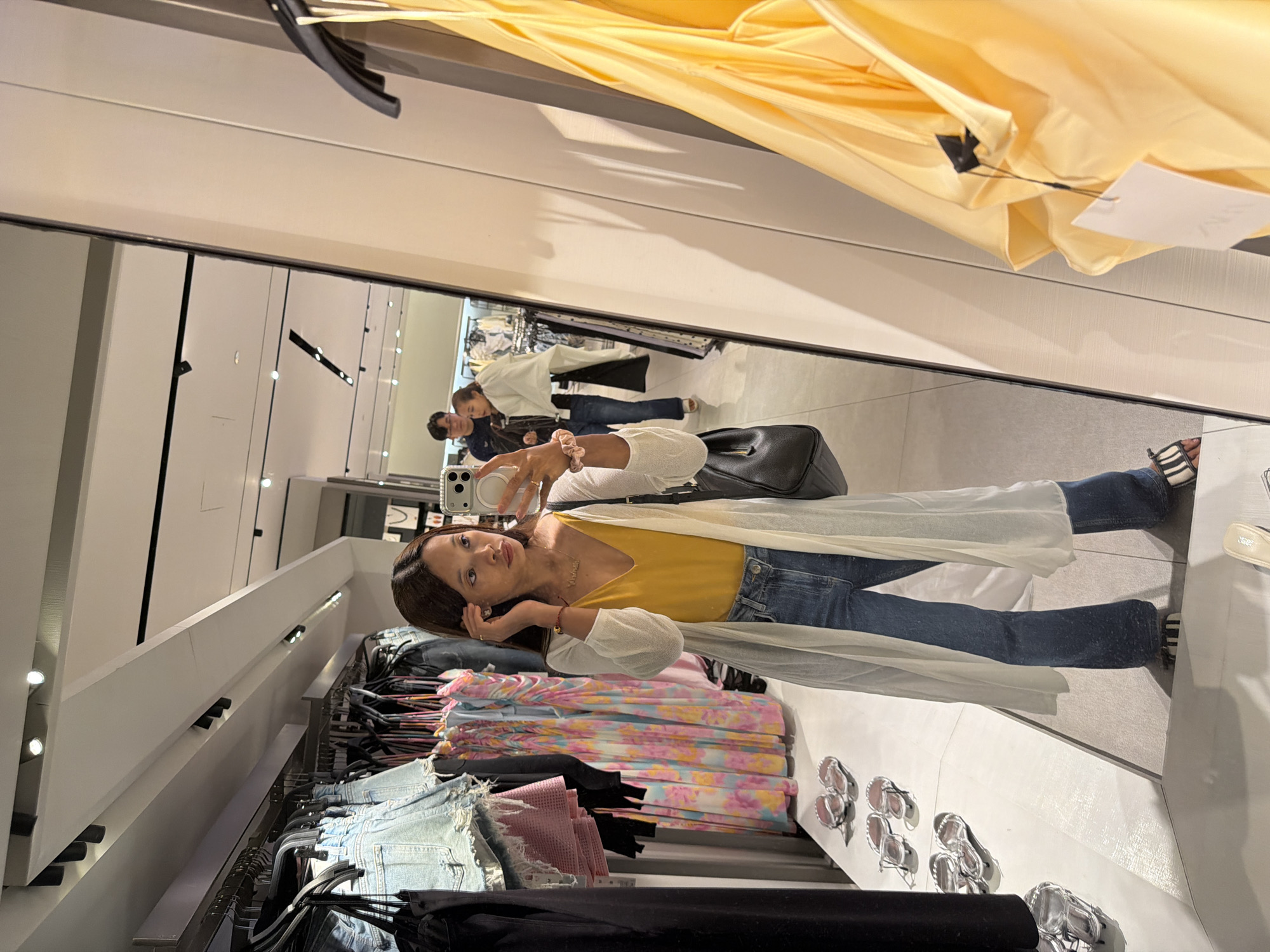 A woman is taking a selfie in front of a mirror inside a Zara store. She is wearing a yellow top, blue jeans, and a white cardigan, and she is holding a smartphone in her right hand. Her hair is dark and appears to be shoulder-length. To her right, there is a rack of clothing. In the background, two people are visible, one wearing a white shirt and dark bottoms, and the other wearing a dark jacket and dark pants. The store setting is suggested by the clothing racks, the mirror, and the lighting. While the location is not explicitly stated, the description indicates the possibility of this image being taken in the city of Kuta, Indonesia, based on the general location.