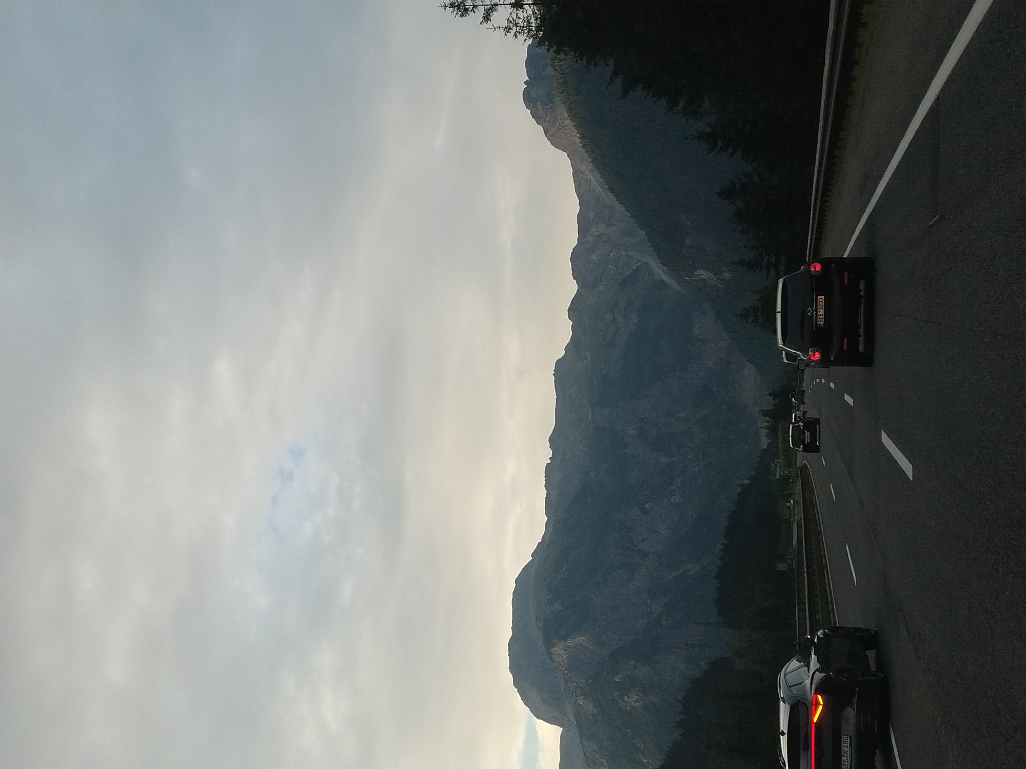 The image depicts a view from a car on a highway in Flachauwinkl, Austria, with a mountain range in the background. The sky is overcast with clouds. Several cars are visible on the highway, including a black SUV in the foreground on the left with its taillights illuminated, and another black SUV further ahead on the right. Trees and mountains form the backdrop, with the mountains appearing dark and rugged, partially obscured by clouds. The highway has multiple lanes with white markings. The scene appears to be during the daytime, possibly late afternoon or early evening, given the lighting conditions.