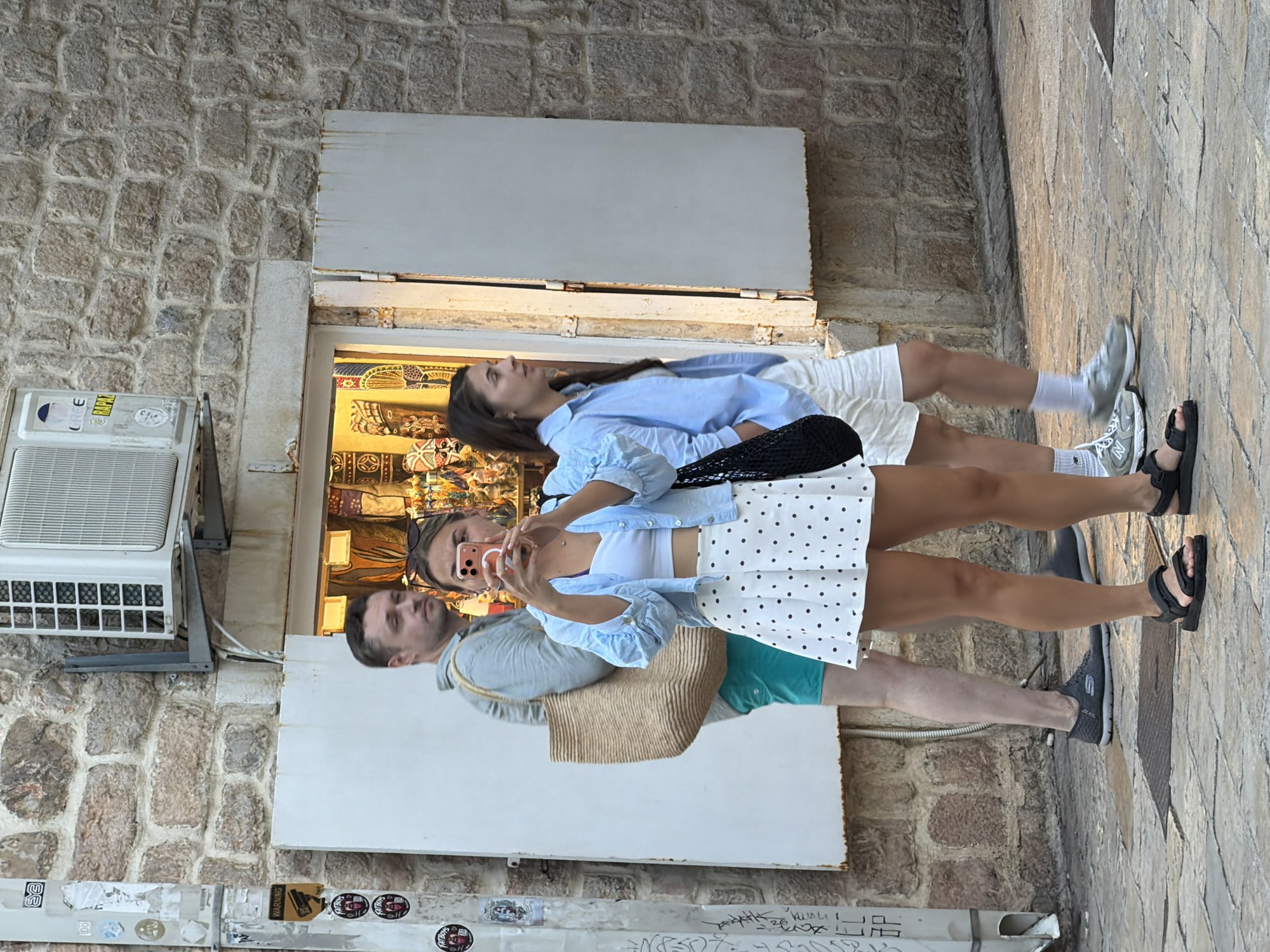 In Budva, Montenegro, on a cobblestone street lined with stone buildings, three individuals are captured in a moment. A woman in the foreground, wearing a white polka-dot skirt and a light blue open-front shirt over a white top, is holding a smartphone in her right hand, seemingly taking a picture or video. Her left hand is obscured. She is wearing black sandals. Behind her, a man with short brown hair and a light grey shirt, carrying a large woven bag over his left shoulder, stands facing left, his turquoise shorts visible. To the right of the woman, a young woman with long dark hair, wearing a light blue button-down shirt over a white outfit and white shorts, is looking to her left, holding a black mesh bag. An air conditioning unit is mounted on the stone wall above a shop window displaying various items, including masks and textiles. The scene is outdoors, with natural light illuminating the area.