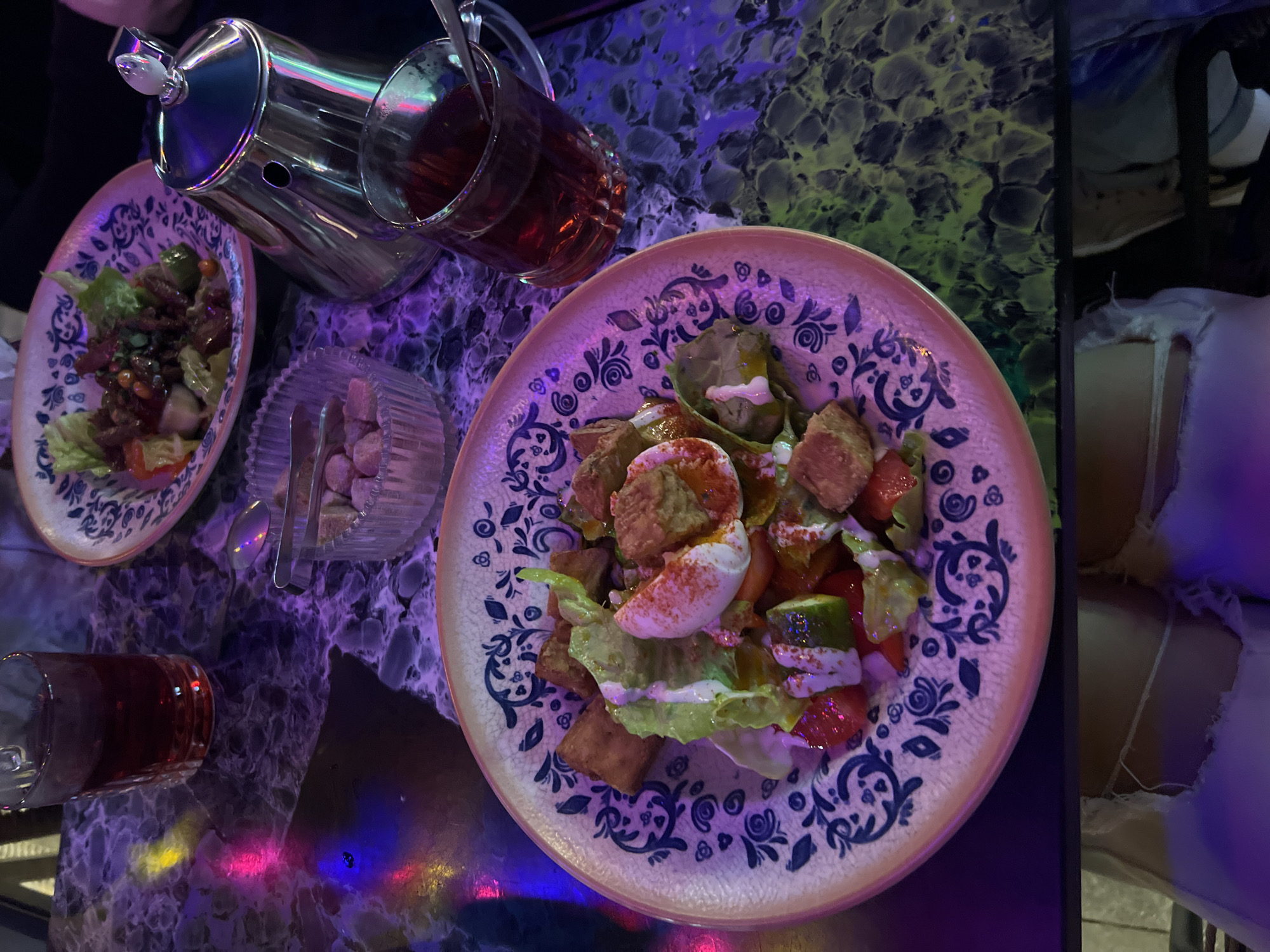 A close-up overhead shot shows two plates of food and drinks on a table with a dark, marbled surface, illuminated by colorful lighting. The plate in the foreground, decorated with a blue and white floral pattern, contains a salad with lettuce, cubed pieces of bread or falafel, chopped tomatoes, cucumbers, and a poached egg. A creamy dressing is drizzled over the salad, and paprika is sprinkled on top. To the left of this plate is a glass of dark red liquid, possibly tea or juice. In the background, another plate with a similar floral pattern holds a salad with lettuce, meat, and vegetables. Next to this plate is a clear glass bowl filled with sugar cubes, and a silver teapot. A glass of the same dark red liquid is positioned to the right of the teapot. The scene appears to be set in a restaurant or cafe. The lighting suggests it is evening or night, with purple and green hues reflecting off the table surface. There is no discernible indication of the specific location in Nizhny Novgorod, Russia, beyond it being an indoor dining establishment.