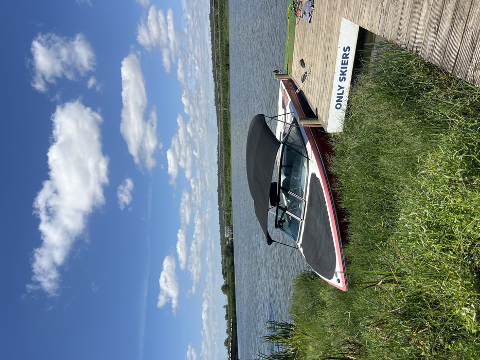 The image displays a red and white pleasure boat, equipped with a black bimini top and a black bow cover, docked at a wooden pier. The boat's bow points towards the overgrown grassy bank on the left, where tall green grass and reeds are abundant. A white sign with blue text reading "ONLY SKIERS" is affixed to the side of the pier facing the water. On the pier's wooden planks, a pair of black flip-flops and a folded grey textile item are visible. The boat is moored in a calm body of water with slight ripples, reflecting the blue sky and scattered white cumulus clouds above. In the mid-ground across the water, green vegetation lines the distant bank, where a large, curved irrigation system and a small, light-colored building are discernible. A bridge structure spans the water in the far left background. The scene is located in Coruche, Portugal, under bright daylight conditions. No individuals are present.