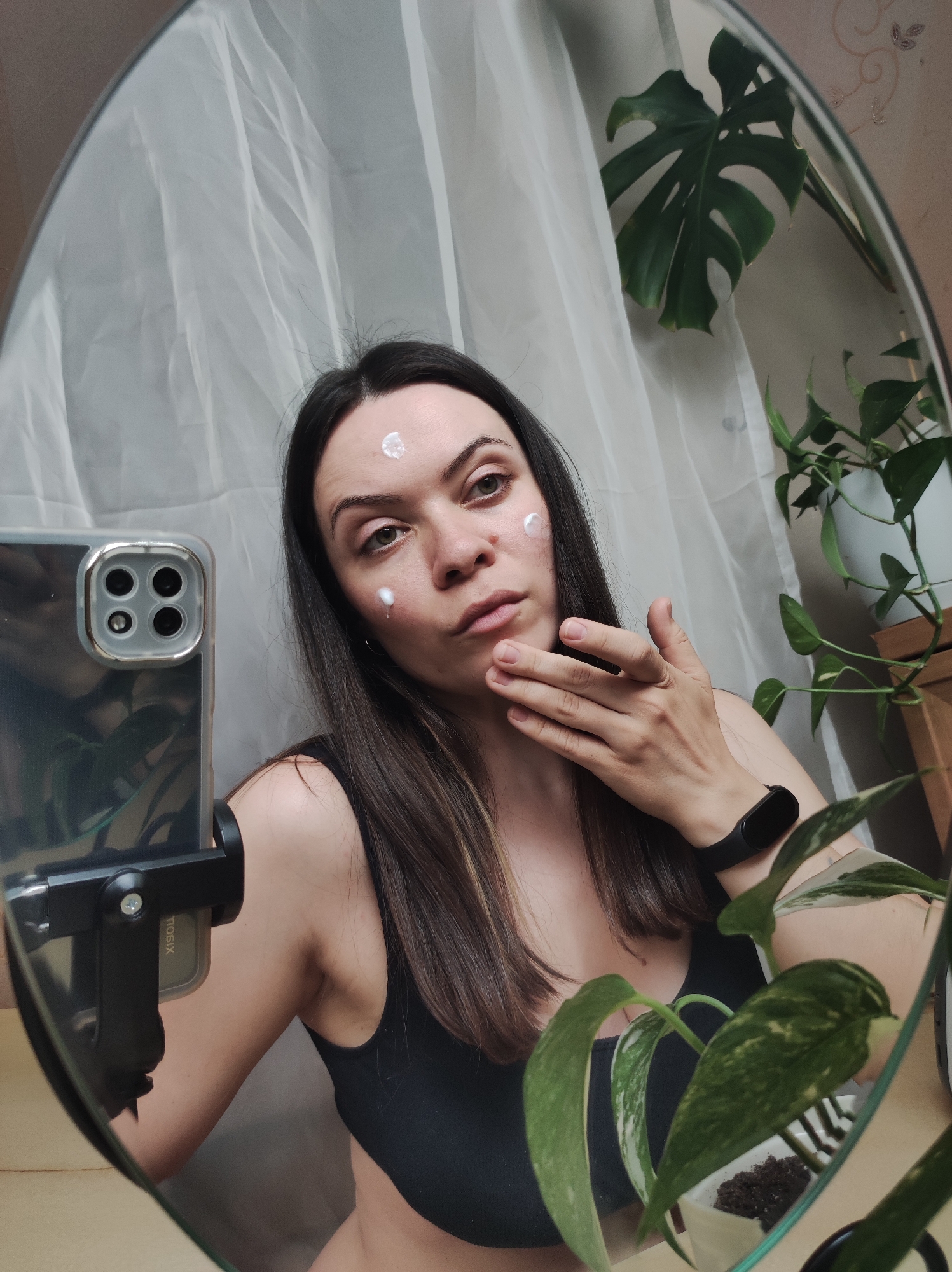 A woman is taking a selfie in front of a mirror. She is wearing a black top and has cream on her forehead and cheek. She is holding her hand up to her face. The mirror is reflecting the woman, a curtain, and some plants.