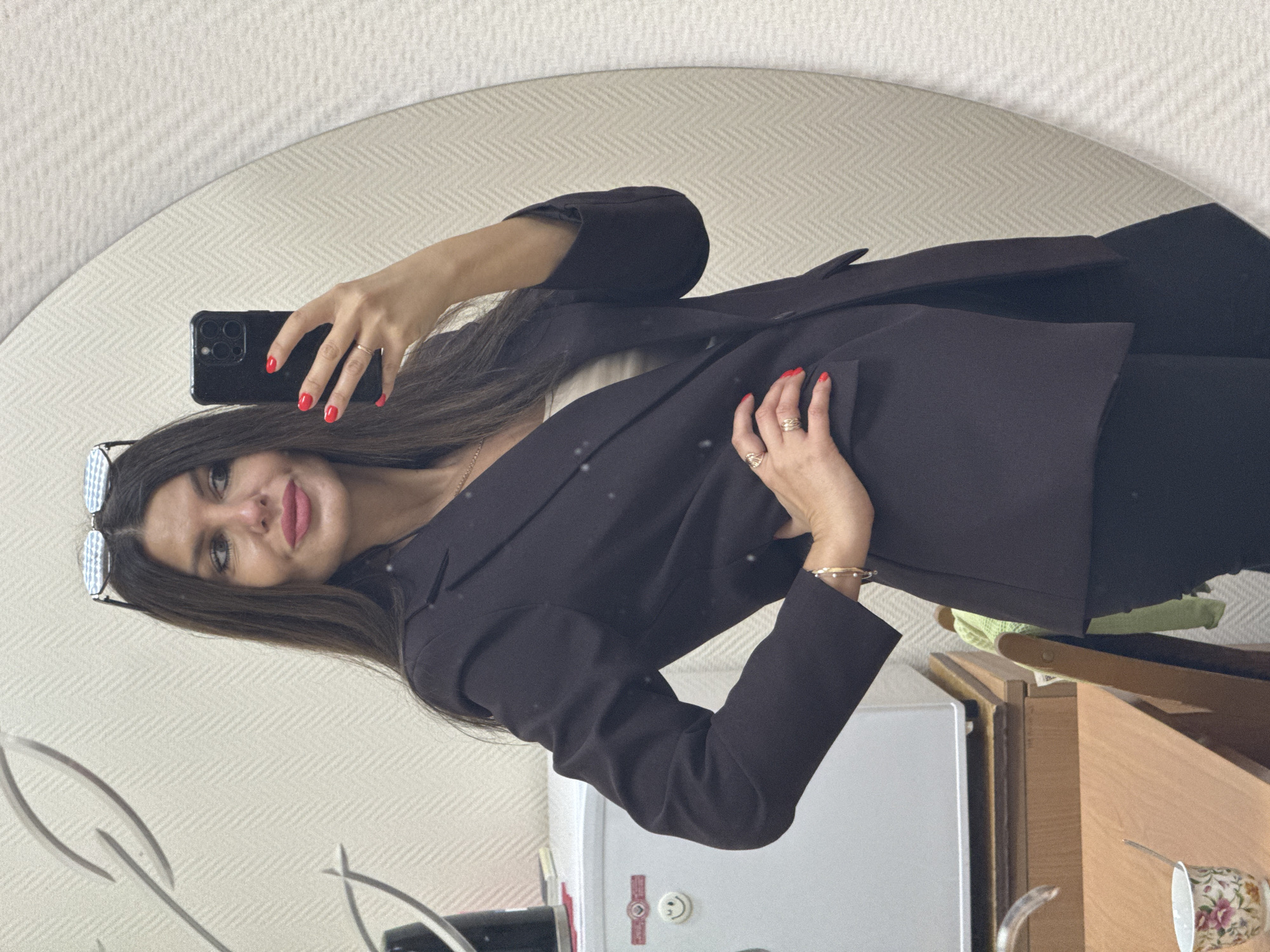 A woman with long, dark hair and red lipstick is taking a selfie in a mirror. She is wearing a dark blazer over a light-colored top and black pants. She has several rings on her left hand, which is resting on her hip. There are glasses perched on top of her head. In the background, a white appliance, possibly a mini-fridge, is visible, along with a wooden desk and a cup with a floral pattern. The wall behind her has a textured, herringbone pattern. The scene appears to be an indoor setting, likely a home or office in the city of Chaikovsky, Russia.