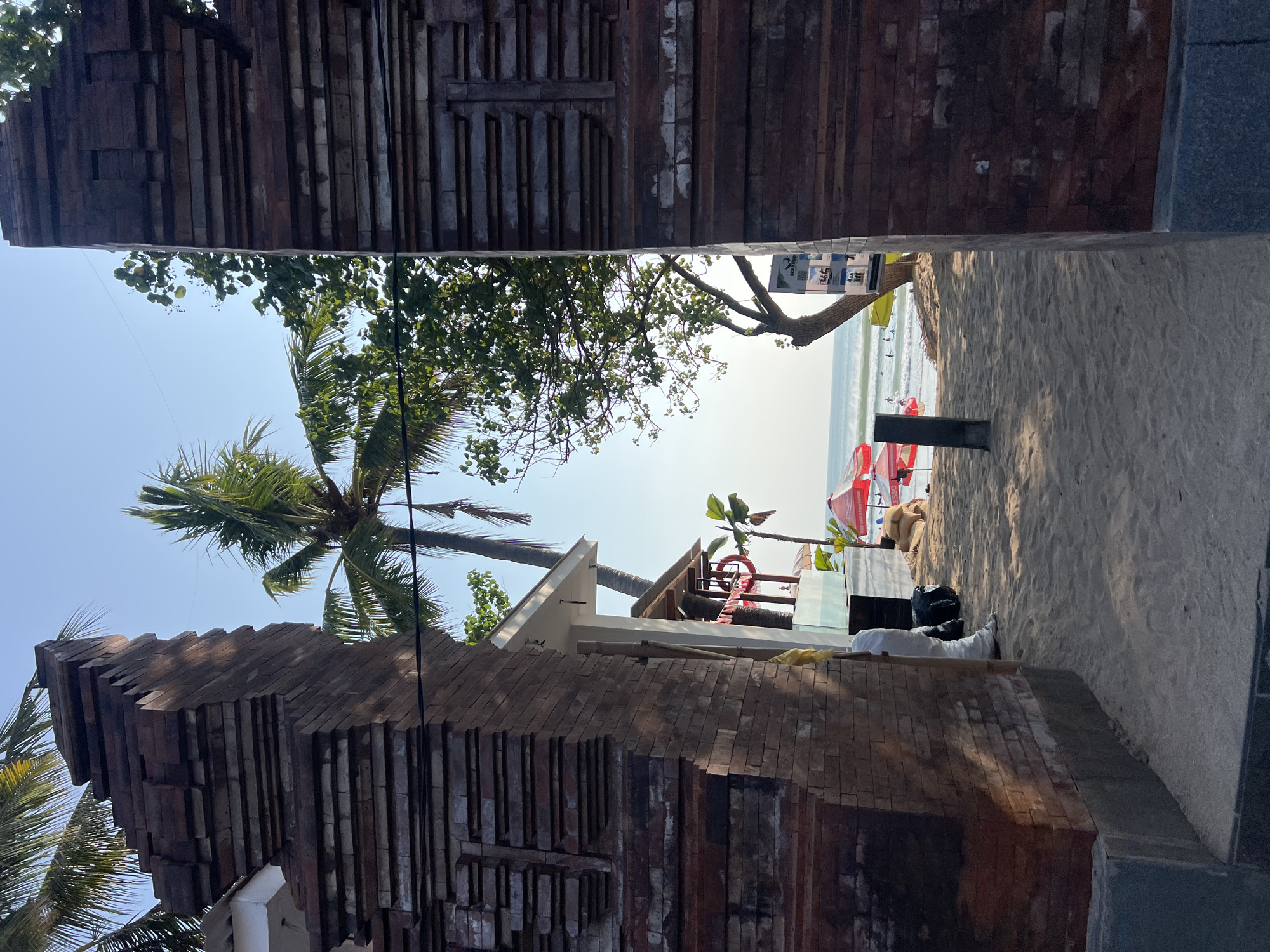 The image shows a view through an ornate brick archway, leading to a sandy beach and the ocean in Kuta, Indonesia. The archway is made of weathered, dark red bricks and features decorative layered stonework. Beyond the arch, palm trees and other foliage are visible against a clear blue sky. The beach is covered in light brown sand and there are several people scattered across it, some near the water and others relaxing under umbrellas. Red and white umbrellas are set up on the sand, and there are bags of what appear to be sand or soil. In the distance, the ocean waves are visible, and a few individuals are in the water, some appearing to be swimming or surfing. A building with a white awning and a red life preserver hangs from it is situated on the left side of the beach. A black trash bag and a yellow object are on the sand near the building. A thin black pole stands in the sand in the foreground, to the right of the center of the frame.