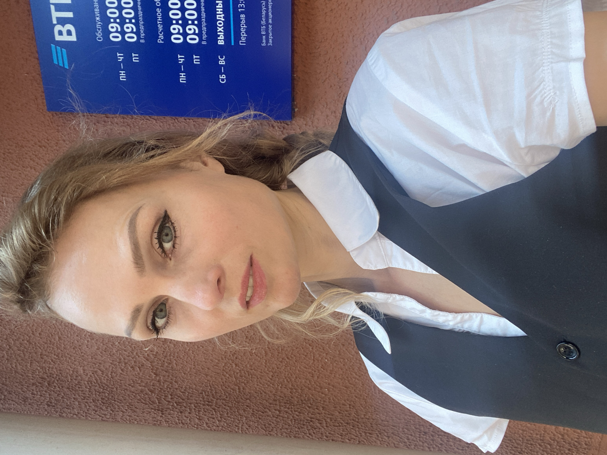 A headshot of a woman in Lida, Belarus. The woman has blonde hair, fair skin, and green eyes. She is wearing a white collared shirt and a black vest. Behind her, a blue sign for VTB Bank is visible. The sign displays operating hours for different services, including "ПН-ЧТ" (Monday-Thursday) and "ПТ" (Friday) from 09:00 to 20:00, and "СБ-ВС" (Saturday-Sunday) as "ВЫХОДНЫЕ" (Weekends). There is also text indicating a break from 13:00 to 13:30 and mentions "Банк ВТБ (Беларусь) Открытое акционерное общество" (VTB Bank (Belarus) Open Joint Stock Company). The background wall is textured and brown.