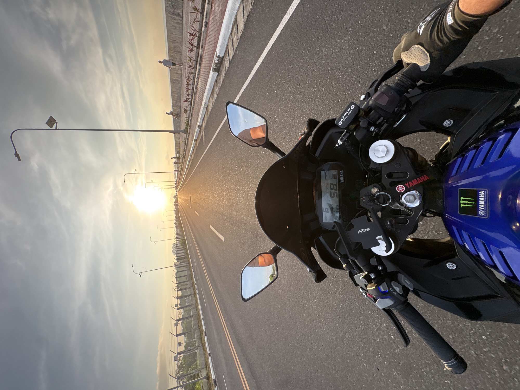 The image is a first-person perspective of a person riding a blue Yamaha motorcycle on a paved road during sunset. The road appears to be part of an elevated highway or bridge, with a concrete barrier on the right side and a barbed wire fence on the left. The sky is a mix of gray clouds and a bright, setting sun casting a warm glow on the scene. In the distance, there are streetlights and several people walking and cycling on a pedestrian walkway along the right side of the road. The motorcycle's dashboard shows a speed of "58". The overall setting suggests a scenic route within Davao City, Philippines, likely along the coastal road or a bridge, given the presence of the sea barrier and the expansive sky.