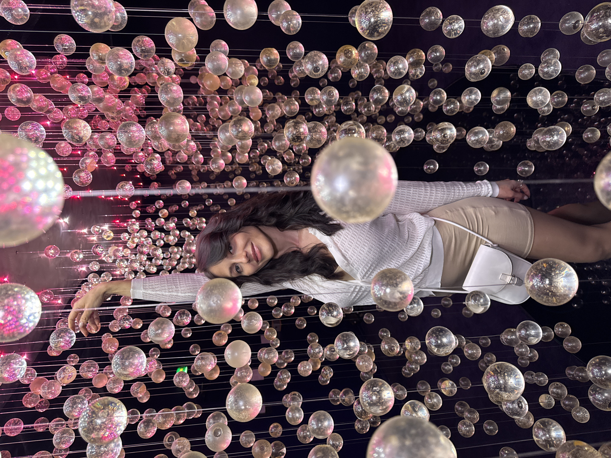A woman stands in front of a dark background adorned with numerous hanging glass orbs of varying sizes. The orbs are lit from behind and from within, creating a shimmering effect with pink and yellow hues. She is wearing a white, long-sleeved, textured top and a light-colored skirt. A small white purse with a strap is slung across her body. Her right arm is raised, with her hand reaching towards the orbs. The scene appears to be an art installation or decorative display. There is no identifiable location in Saint Petersburg, Russia.