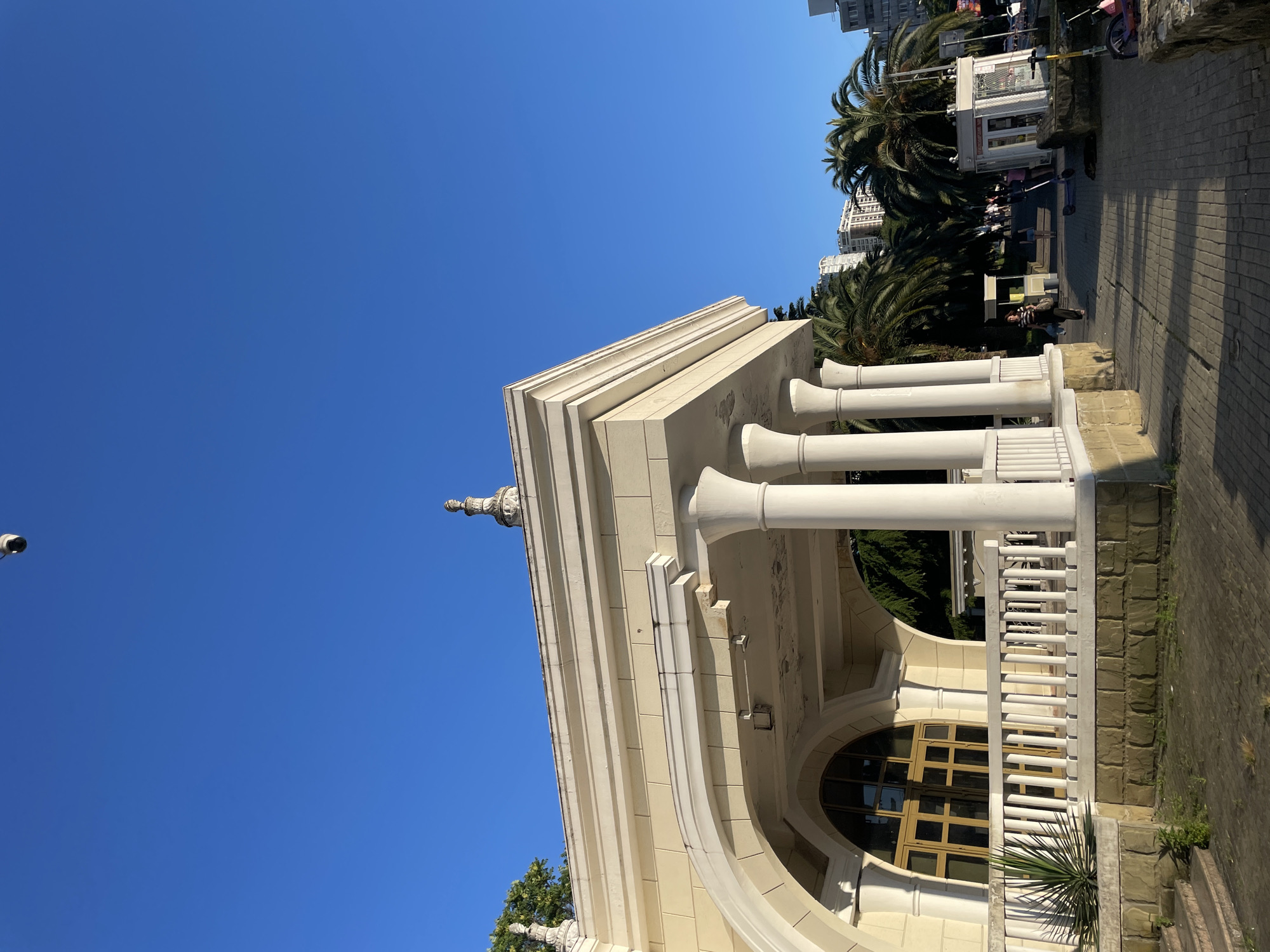 The image depicts a sunny day in Sochi, Russia, specifically near the port area. In the foreground, a light-colored building with classical architectural elements stands. It features a covered porch supported by white columns and a white railing. Behind the building, palm trees and modern apartment buildings are visible, indicative of a coastal resort city. A paved walkway runs alongside the building, with a few people walking and what appear to be small kiosks or stands. A security camera is mounted high on the building, and another is visible on a pole in the distance. The sky is a clear, vibrant blue.
