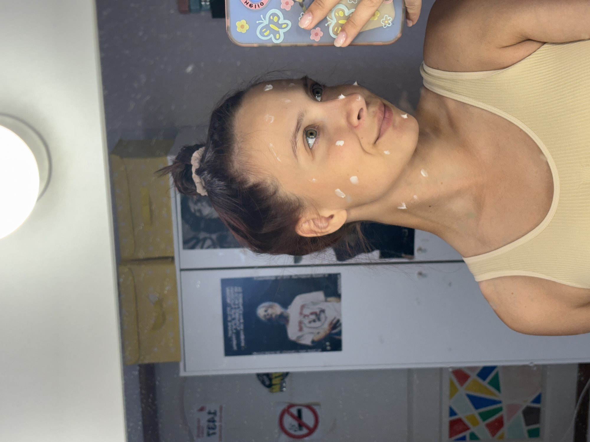 A young woman with light skin and green eyes is seen in a selfie-style image, likely taken in a bathroom. She has dark hair pulled back in a bun. Small dots of what appears to be cream or lotion are applied to her face and neck. She is wearing a light-colored ribbed tank top. She is holding a mobile phone with a colorful case.
The background is a bathroom mirror reflecting the scene behind the photographer. This includes a white cabinet with yellow patterned storage bins, a framed poster of a person, and a no-smoking sign, along with a colorful mosaic tile section. The location of Прикамский, Russia is not suggested by any element in the image.