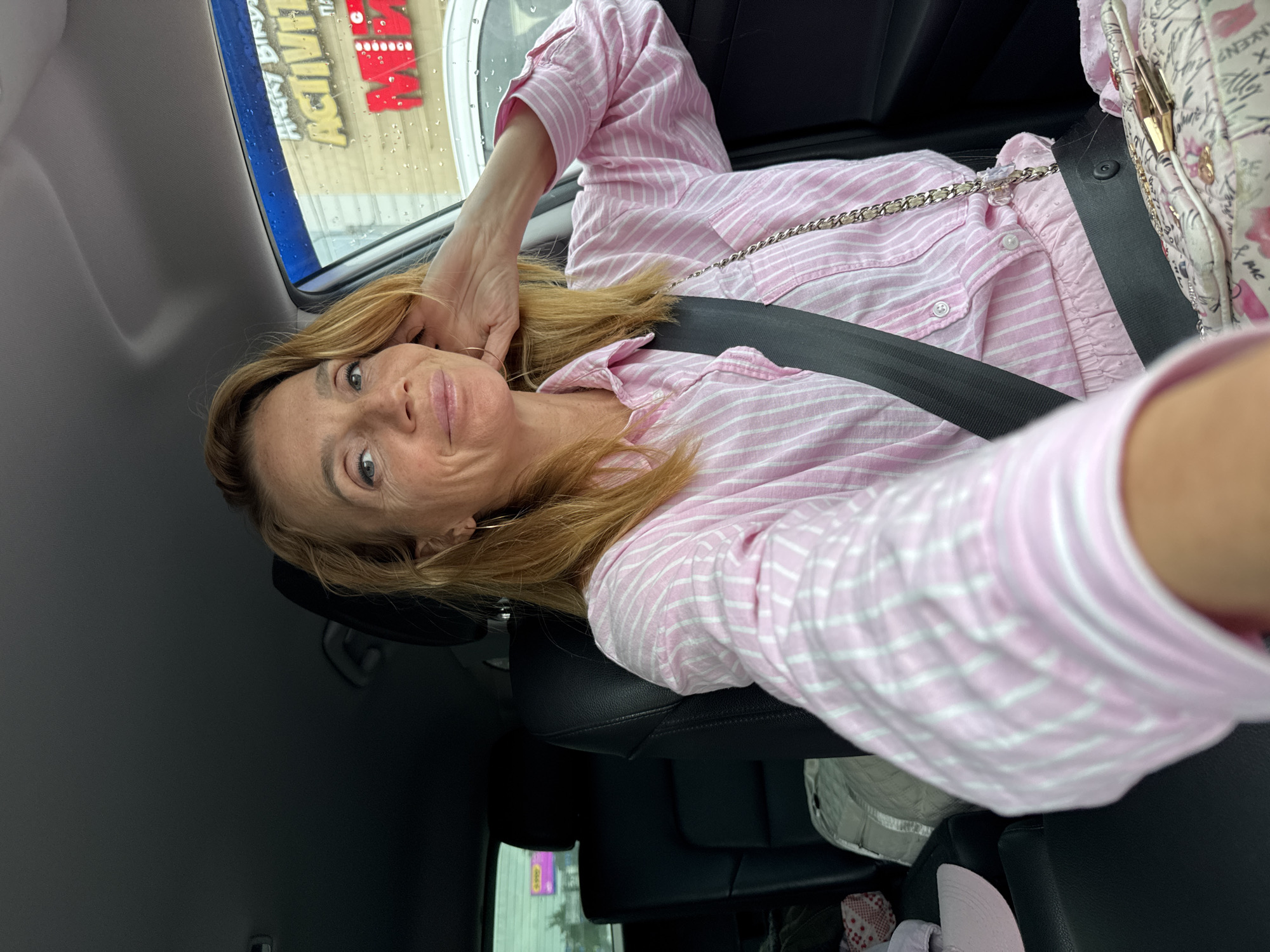 A woman with blonde hair and light skin is sitting in the driver's seat of a car. She is wearing a pink and white striped button-down shirt and a seatbelt. Her right hand is resting on her head. Outside the car window, a building with a sign that reads "ANGRY BIRDS ACTIVITY PARK" is visible. The weather appears to be rainy. The interior of the car is dark, with black leather seats.