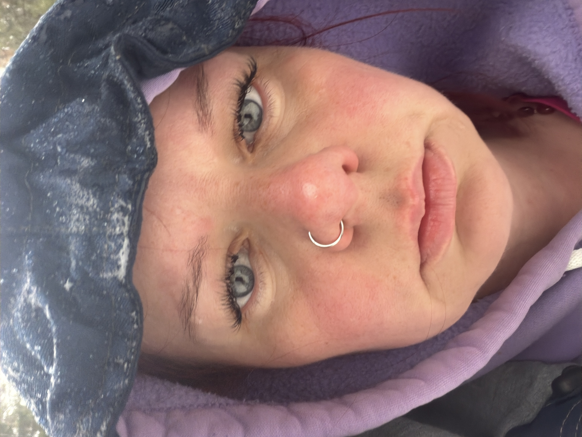 A close-up, tilted portrait depicts a fair-skinned individual with a silver nose ring. The person has gray eyes, visible eyelashes, and reddish hair strands. The person is wearing a lilac-colored hooded sweatshirt. Part of a dark blue, possibly denim, hooded jacket, with what appears to be frost or white speckles, is visible covering the top left of the frame. The individual's face appears red or flushed. The image provides no evidence to suggest a location in Chaikovsky, Russia.
