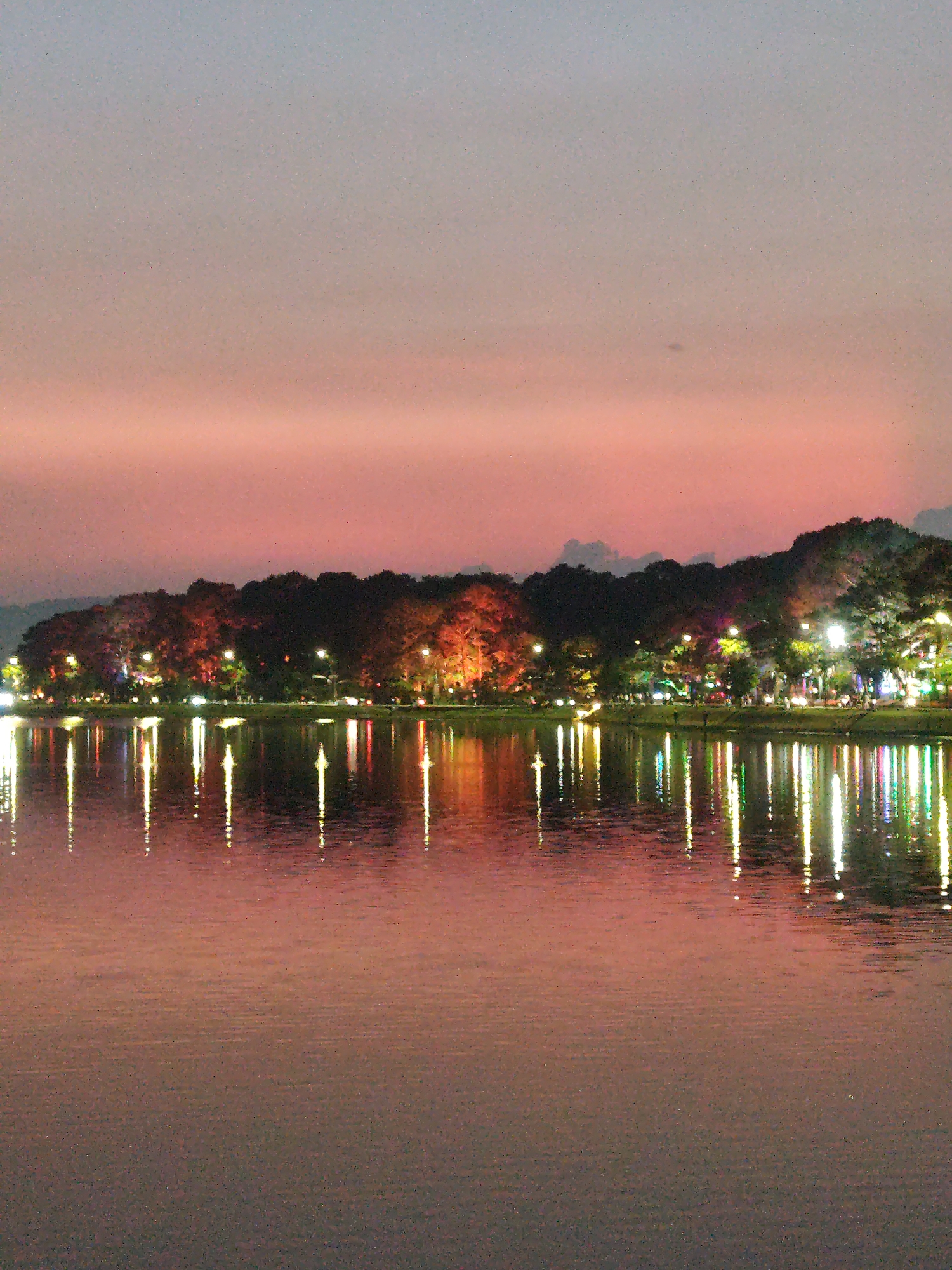 The image depicts a tranquil lake scene in Da Lat, Vietnam at dusk. The sky transitions from a soft pink to a muted gray. The far shore is lined with trees and illuminated by streetlights and building lights, creating a warm, diffuse glow. The lights are reflected in the still water, forming vertical lines of bright colors. The water's surface is calm with subtle ripples near the foreground. No people are clearly visible, but the presence of lights suggests human activity along the shore. The environment suggests a park or recreational area adjacent to a lake within a city.