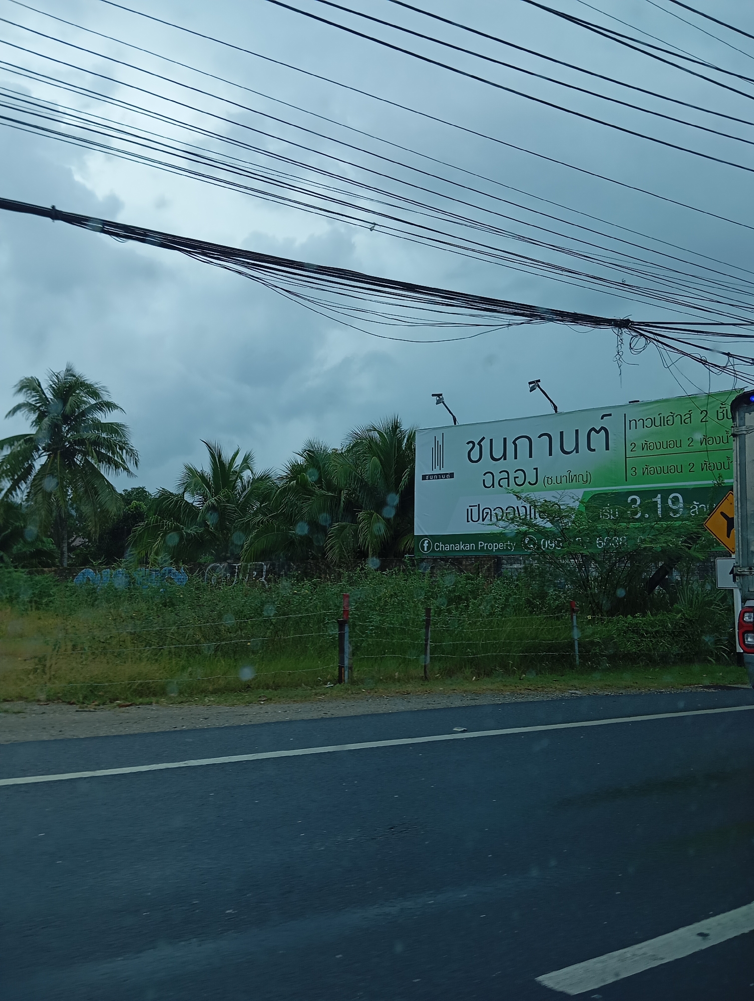 The image shows a billboard on the side of a road. The billboard advertises "CHANA KAN CHLONG (Na Yai) Houses from 2.19 million". The billboard has text in Thai and an image of houses. There are also power lines overhead and palm trees in the background. The road is wet, suggesting it may have been raining. The overall scene depicts an outdoor advertisement in a tropical or subtropical environment.