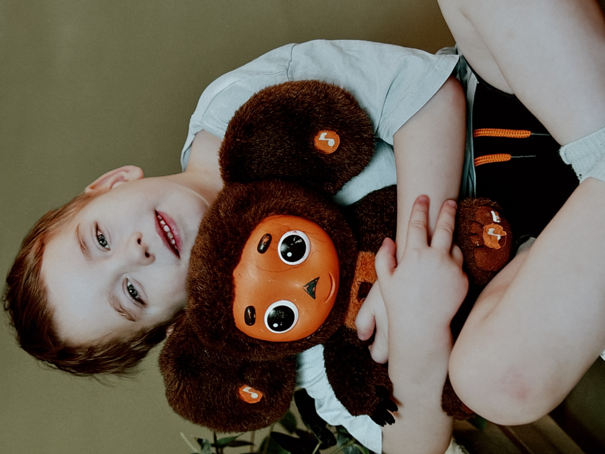A young child lies supine, cradling a brown Cheburashka plush toy. The child has fair skin, reddish-brown hair, and blue eyes. They are wearing a light-colored t-shirt and dark shorts with orange accents. The Cheburashka toy is predominantly brown with a large orange face and ears. The background is a muted green. There is no evidence to support the claim that the image was taken in Chaikovsky, Russia.