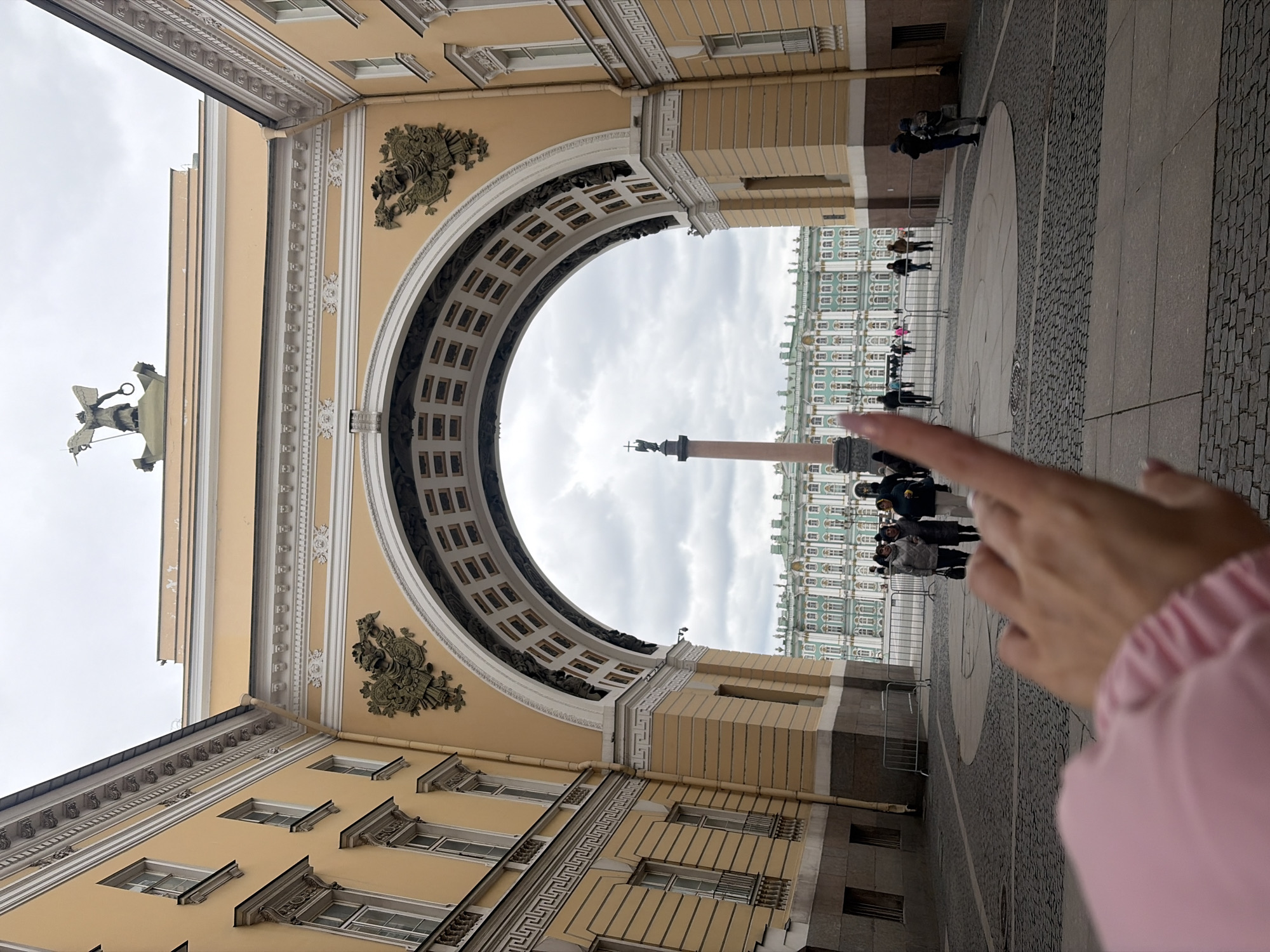 The image presents a grand architectural vista of Palace Square in Saint Petersburg, Russia. The scene is dominated by the monumental archway of the General Staff Building, which spans across the foreground. This light yellow building features intricate white neoclassical detailing, including cornices, pilasters, and elaborate relief sculptures. High above the left side of the arch (when viewed upright), a bronze statue of a chariot, known as the Chariot of Glory featuring Nike, is visible against the overcast sky.
Through the arch, the expansive cobblestone and paved square stretches into the distance. In the center of the square stands the towering Alexander Column, a monolithic red granite column topped with a statue of an angel holding a cross. Beyond the column, the iconic light green and white Baroque facade of the Winter Palace, home to the State Hermitage Museum, lines the far side of the square.
Numerous people are visible scattered across the square, appearing to be tourists or visitors exploring the historical site. Metal crowd control barriers are positioned in parts of the plaza. In the bottom right foreground, a human hand with a pink sleeve is prominently featured, pointing directly towards the Alexander Column.
The weather appears to be overcast, with a uniform grey sky suggesting a cloudy day. The time of day is not precisely discernible but is clearly daylight. No discernible text is visible on any of the buildings or monuments. The overall impression is one of historical grandeur and architectural splendor.