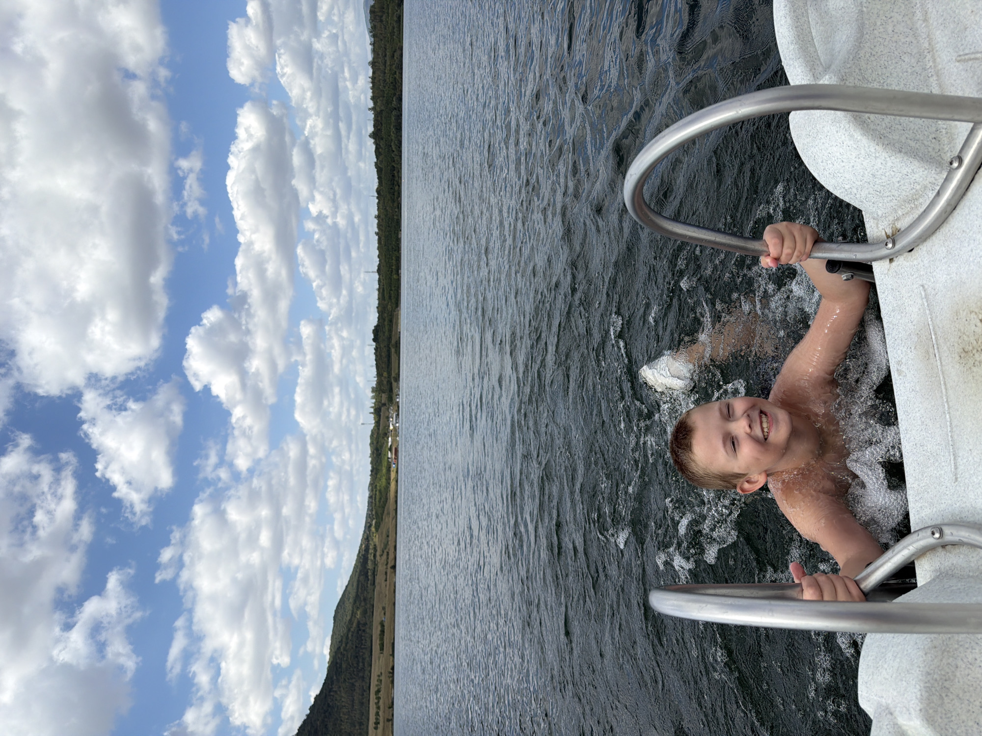 In this image, a young boy is being photographed while in a body of water, holding onto a metal railing. He appears to be swimming or playing in the water, as his body is partially submerged and he is smiling broadly at the camera. The water is dark and choppy, suggesting movement. The boy is light-skinned with short, light brown hair. He is wearing white sneakers, which are visible at the edge of the frame.
The setting is a lake or large body of water, with a shoreline visible in the background. The shoreline is lined with trees, forming a dark green strip against the landscape. Above the trees, a hillside is visible, covered with vegetation that appears dry and yellowed in places, suggesting a summer or early autumn setting. The sky is a vibrant blue, filled with fluffy white clouds. The lighting suggests it is daytime, likely afternoon, with the sun casting a soft glow.
The overall mood is joyful and carefree, as conveyed by the boy's happy expression and the recreational activity. There is no visible text in the image. The location context provided is "Most, Czechia."