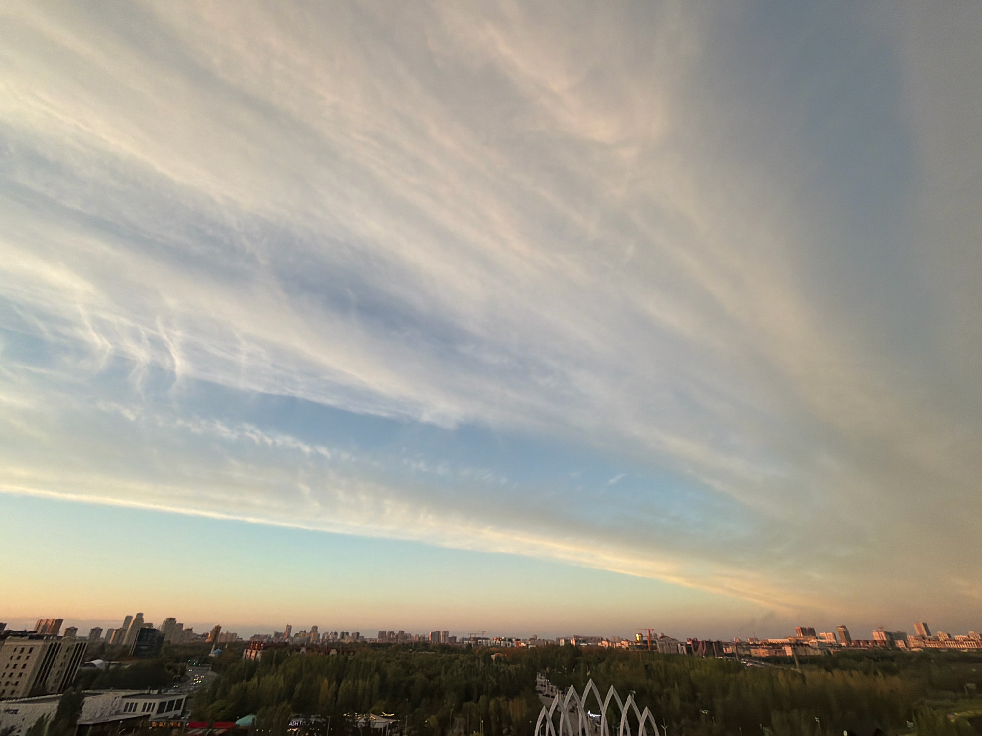 The image depicts a panoramic view of Astana, Kazakhstan, during what appears to be sunset or sunrise. The sky is filled with wispy, elongated clouds that stretch across the frame, transitioning from a pale blue near the horizon to a muted gray at the top. The clouds exhibit subtle pink and yellow hues, indicating the low angle of the sun.
Below the sky, the cityscape of Astana is visible. Tall buildings, some with distinctive modern architecture, are scattered across the horizon. A dense expanse of green trees forms a significant portion of the mid-ground, suggesting a large park or forested area within the city. In the foreground, parts of a white, arched structure, possibly an architectural element of a park or public space, are visible on the right side. The overall impression is of an urban landscape bathed in soft, natural light.