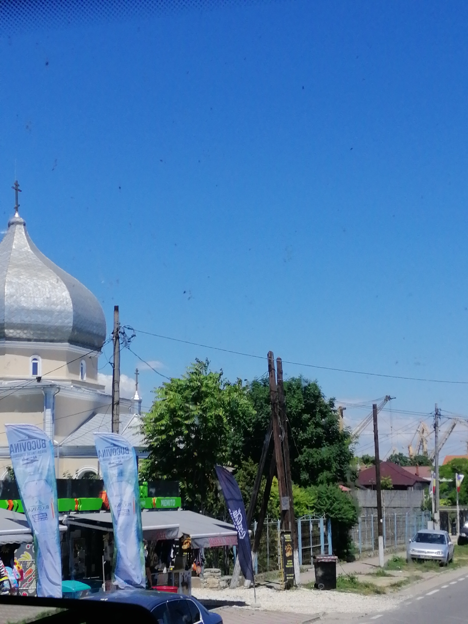 The image depicts a street scene in 2 Mai, Romania, on a bright, clear day. A prominent silver-domed church with a cross on top is visible on the left side of the frame. In front of the church, there are several market stalls with awnings. Two large blue flags bearing text and graphics are displayed near the stalls. A dark blue car is parked in the foreground, partially obscuring the view of the stalls and flags. In the background, there are trees, some houses with red roofs, and industrial cranes. A silver car is parked on the right side of the street. The street is paved, and there is a sidewalk lined with gravel and some vegetation. Electrical poles with wires are also visible. The sky is a vibrant blue with a few scattered dark spots, possibly dust or small insects on the camera lens.