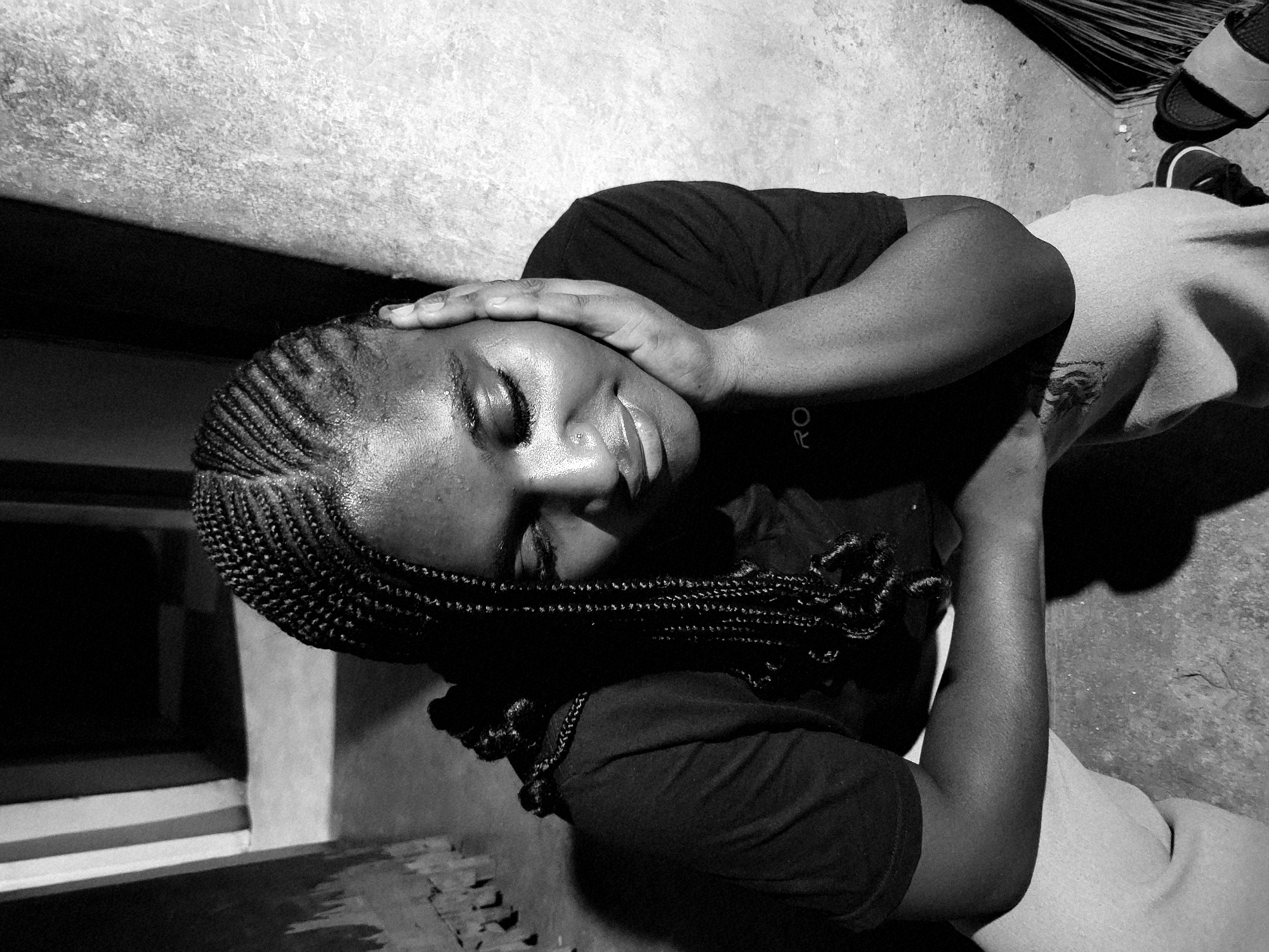 The black and white image depicts a person with dark skin and braided hair, lying on what appears to be a concrete surface, possibly a porch or patio. The individual is wearing a dark t-shirt and light-colored pants. Their eyes are closed, and one hand is resting on their forehead. The setting includes a window with horizontal slats, a broom leaning against the wall, and a pair of shoes. The location cannot be forensically confirmed as the city of Jos, Nigeria, without additional data.