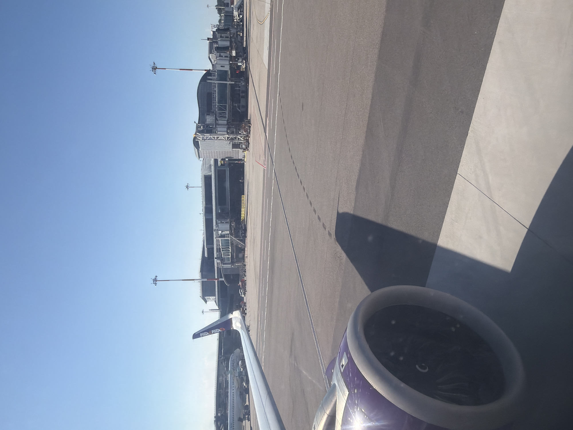 The image was taken from the interior of an aircraft, looking out a window at an airport. In the foreground, the engine nacelle of the aircraft is visible, painted in shades of purple and gray, with a bright sheen from sunlight. The background shows an airport terminal building with jet bridges connecting to aircraft. Several other airplanes are parked on the tarmac, along with service vehicles and lighting poles. The sky is clear and blue. The scene is of an airport environment in the city of Ferno, Italy, likely Malpensa Airport, a major international airport serving Milan.