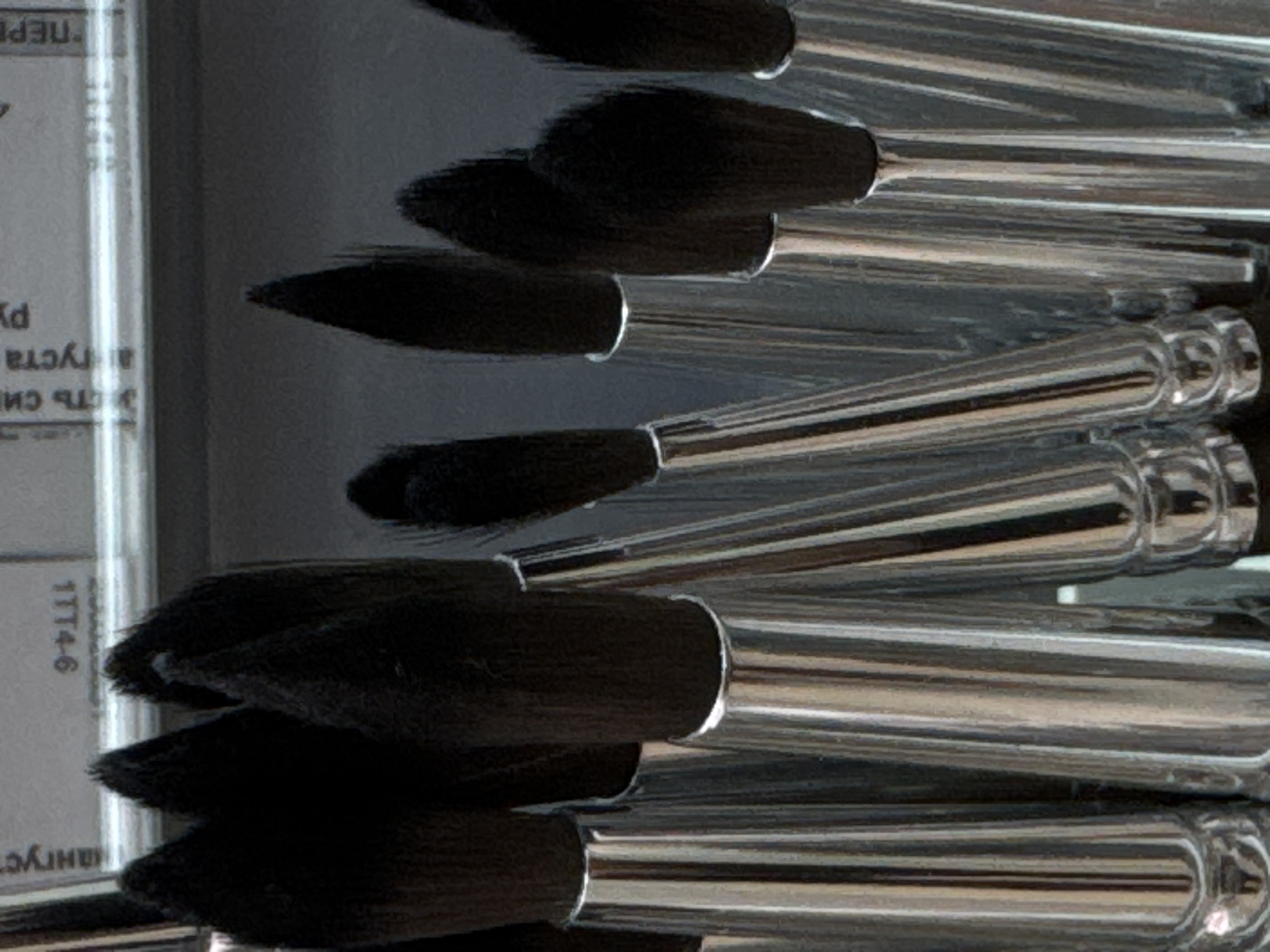 A close-up, low-angle shot shows a collection of paintbrushes with black bristles and shiny silver ferrules. The brushes are arranged in a group, with some overlapping. The background is blurred, but appears to be a surface with some text and possibly a ruler. The lighting is somewhat dim, creating a moody atmosphere. The image does not provide any information about the location in Moscow, Russia, nor does it depict any people, actions, or interactions.