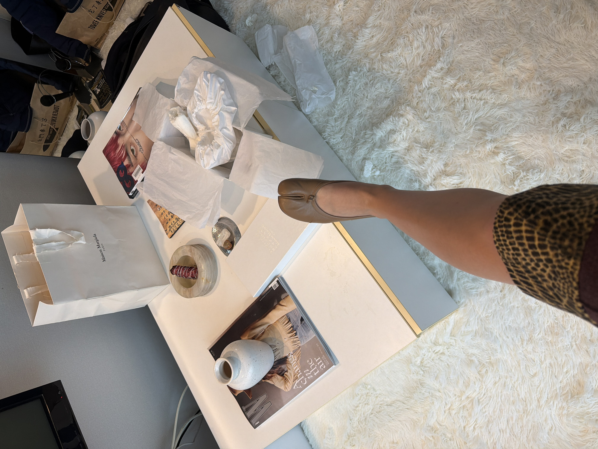 This overhead shot captures an indoor setting, likely a casual living room or hotel room, characterized by a plush, white shag rug covering the floor. A person's bare leg and foot, adorned with a single brown Maison Margiela Tabi shoe, extends into the frame from the right, resting near a modern white coffee table with gold trim. A portion of leopard-print clothing is visible at the top right.
The white coffee table is central to the image, cluttered with items suggesting recent shopping and unboxing. A white paper shopping bag with "Maison Margiela PARIS" printed on it stands on the left side of the table. Nearby, numerous crumpled sheets of white tissue paper are scattered, some appearing to be shoe covers or packaging. Among these papers is a magazine or card featuring a close-up of a person with red hair and detailed eye makeup, with text "CAT" and the number "8" visible.
Further along the table, a circular, light-colored marble dish holds a bundle of what appears to be dried herbs, possibly sage for smudging. A small, speckled white ceramic vase rests on an open "W" magazine, which prominently displays the title "A change in the air." In the background, several brown paper bags are visible, one clearly marked with "TOWER KNIVES" and the Japanese text "包丁専門" (knife specialty). A black television screen is partially visible in the bottom-left corner, against a light gray wall.
The scene suggests a relaxed atmosphere, perhaps after a shopping excursion in Seoul, with new items being unboxed and displayed. The visible text elements provide brand cues for high-end fashion and specialty goods.