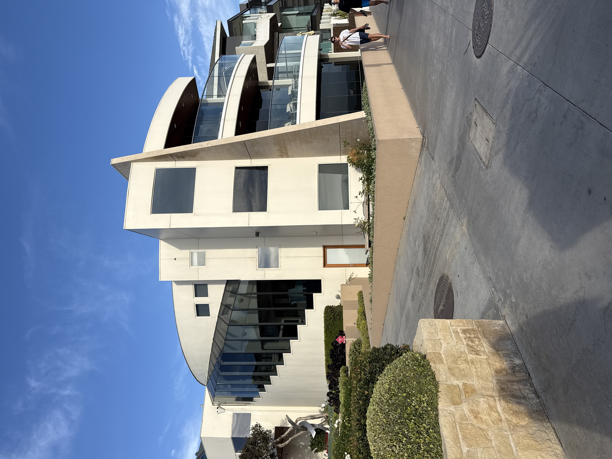 The image depicts a modern, multi-story residential building with a curved facade and several balconies. The building is constructed with cream-colored stucco and features large windows, including a glass-enclosed staircase. The ground level has a tan-colored base. In front of the building, there is a concrete walkway, a stone retaining wall, and manicured landscaping with bushes and flowers. Two individuals are walking on the walkway. The sky above is blue with scattered white clouds. The scene appears to be a residential area in Manhattan Beach, California, likely near the coast, given the architectural style and the presence of a walkway that could be a beach path.
