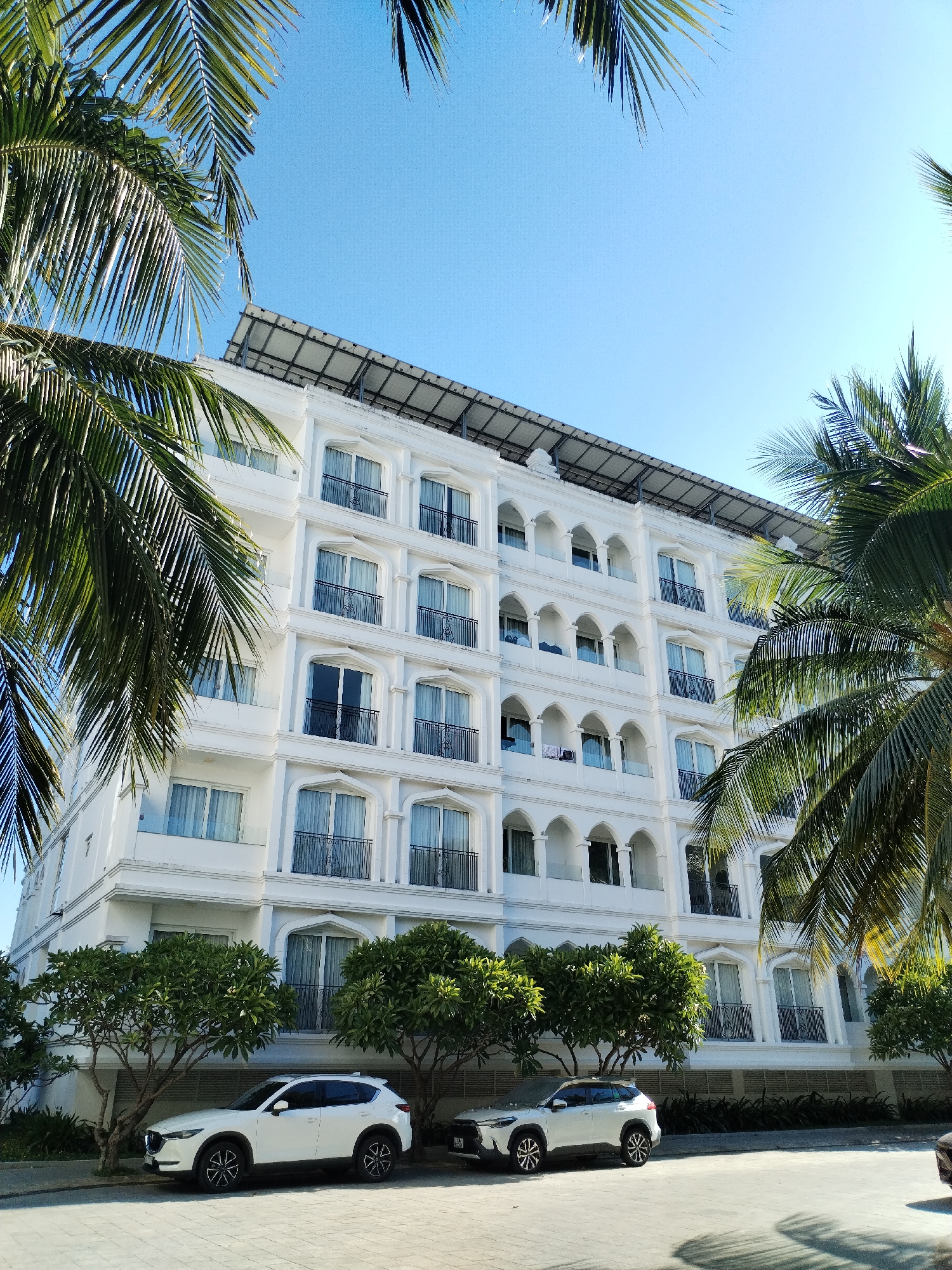 This image shows a white, multi-story hotel with several balconies and arched windows, framed by palm trees. Two white SUVs are parked in front of the hotel on a paved surface. The sky is clear and blue. The location appears to be in a tropical or subtropical region, potentially near a beach or resort area. The architecture suggests a modern, resort-style hotel. The scene is bright and sunny, indicating it was taken during the daytime.