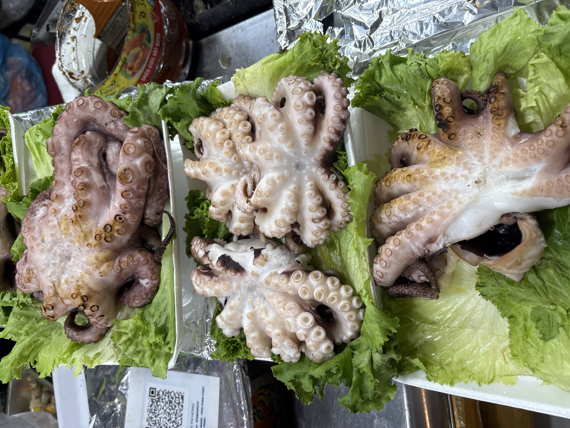 The image captures an overhead view of three portions of cooked octopus, each meticulously arranged on a bed of vibrant green lettuce leaves within white, possibly disposable, trays. The leftmost octopus exhibits a reddish-brown color with distinct suction cups, some displaying darker, charred marks, suggesting grilling or broiling. The central octopus is lighter, appearing white with pinkish tones, its tentacles coiled to reveal an array of suction cups. The octopus on the right also features some browned areas on its suction cups and tentacles.
In the background, elements of a food preparation or serving area are visible, including crumpled aluminum foil. A clear plastic container with an orange label is partially seen in the upper left, bearing the Vietnamese text "CÀNG GION HÀN QUỐC," which translates to "Korean Crunchy Legs" or "Korean Crispy Tendons," likely for a sauce or ingredient. Below the leftmost food tray, a white label with a QR code is visible. Text on this label includes "TRAN THI HONG," a phone number "SĐ T: 00588199777," and partial address or name details such as "NG : PSD PHAN CHAU" and "TRỊNH." The scene indicates prepared seafood, likely from a street vendor or market stall, ready for consumption in a casual setting.