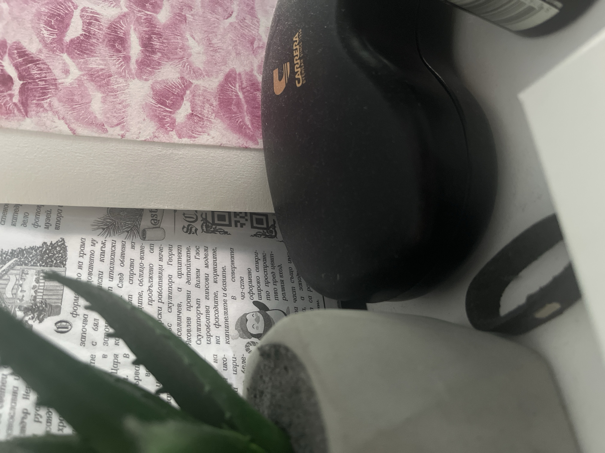This media file presents an angled, close-up view of a cluttered surface, likely a desk or shelf, in an indoor setting in Pokrovnik, Bulgaria. The image features several distinct objects.
In the upper left, a background item displays a repeating pattern of vivid pink lipstick kisses on a white surface, possibly decorative paper or fabric. Dominating the upper right and center is a dusty, black, oblong object, which appears to be an eyewear case. Prominently featured on this item is the gold-colored text "CARRERA EYEWEAR SINCE 1956".
Spanning the lower left and center, an open book or newspaper is visible, displaying Cyrillic text and some illustrations, including what looks like a building, pine branches, and a small figure resembling a penguin. Visible Cyrillic phrases include "по на храма" (of the temple), "скулптора Георги" (sculptor Georgi), "архитект Яковлев" (architect Yakovlev), "Вилем Глос" (Vilem Glos), "гипсови модели" (plaster models), "фасадите" (facades), "корнизите" (cornices), "капителите" (capitals), "базите" (bases), and "северната ча-сте" (northern part).
In the bottom left foreground, a green succulent plant is partially visible, growing from a grey-textured pot. To the far right, a curved dark object and a white surface suggest additional items or architectural features. The overall scene conveys a casual, everyday atmosphere, with the dust on the Carrera case suggesting it might not be a frequently handled item. No people are present, and no specific time of day or weather conditions can be inferred.