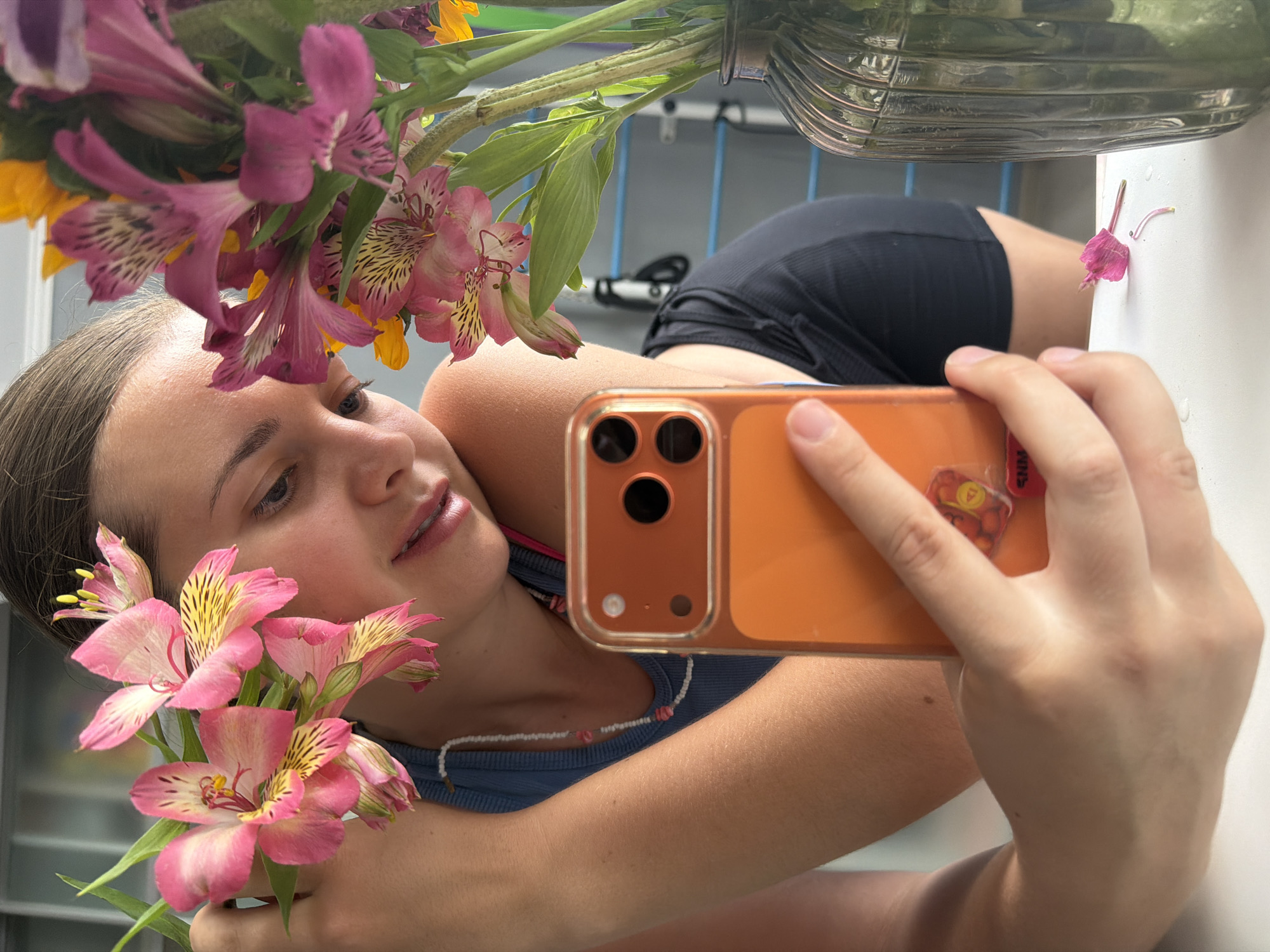 The scene is located in Sochi, Russia.
A young female is depicted in a medium shot, engaged in self-photography or viewing her reflection, holding an orange-cased smartphone in her right hand. Her head is tilted slightly down and to the right, with her gaze directed towards the phone. She has dark hair pulled back, a clear complexion, and exhibits braces on her lower dental arch. She is wearing a light blue ribbed tank top over what appears to be a dark sports bra or similar garment, and a white beaded necklace with small pink elements.
To her left, she holds a small bouquet of pink Alstroemeria flowers, characterized by yellow and dark brown streaking on the petals, positioned near her face. To her right, a dark, ribbed glass vase contains a larger arrangement of similar pink and purple Alstroemeria flowers, interspersed with yellow flowers, resting on a white surface. Two detached pink petals are visible on this white surface in the bottom right foreground.
The smartphone features a multi-lens camera system on its back. The background is blurred, revealing light-colored walls or surfaces and distinct horizontal blue bars, suggesting an indoor setting with possibly gym equipment or specific furniture. The overall lighting is bright and diffuse.