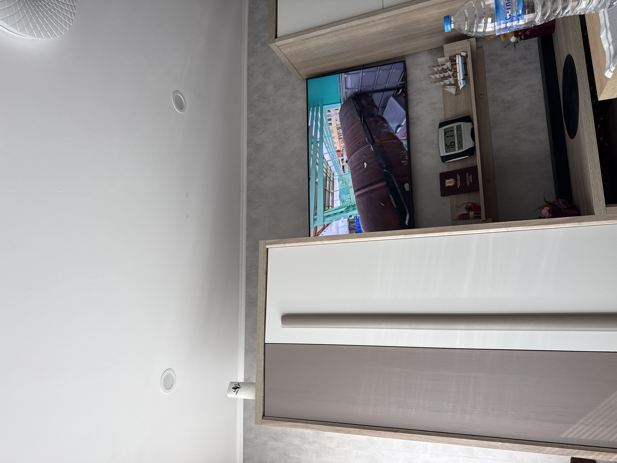 Here is a description of the image:
The image shows an interior scene with a TV mounted on a wall unit. The TV displays a view from a high structure, likely a bridge or tower. A bottle of water and some decorative objects sit on the shelf above the TV. The wall unit has white and grey drawers with a sleek, modern design. The ceiling is white with recessed lighting. The location appears to be indoors. The image does not contain any information that connects it to Lovech, Bulgaria.
