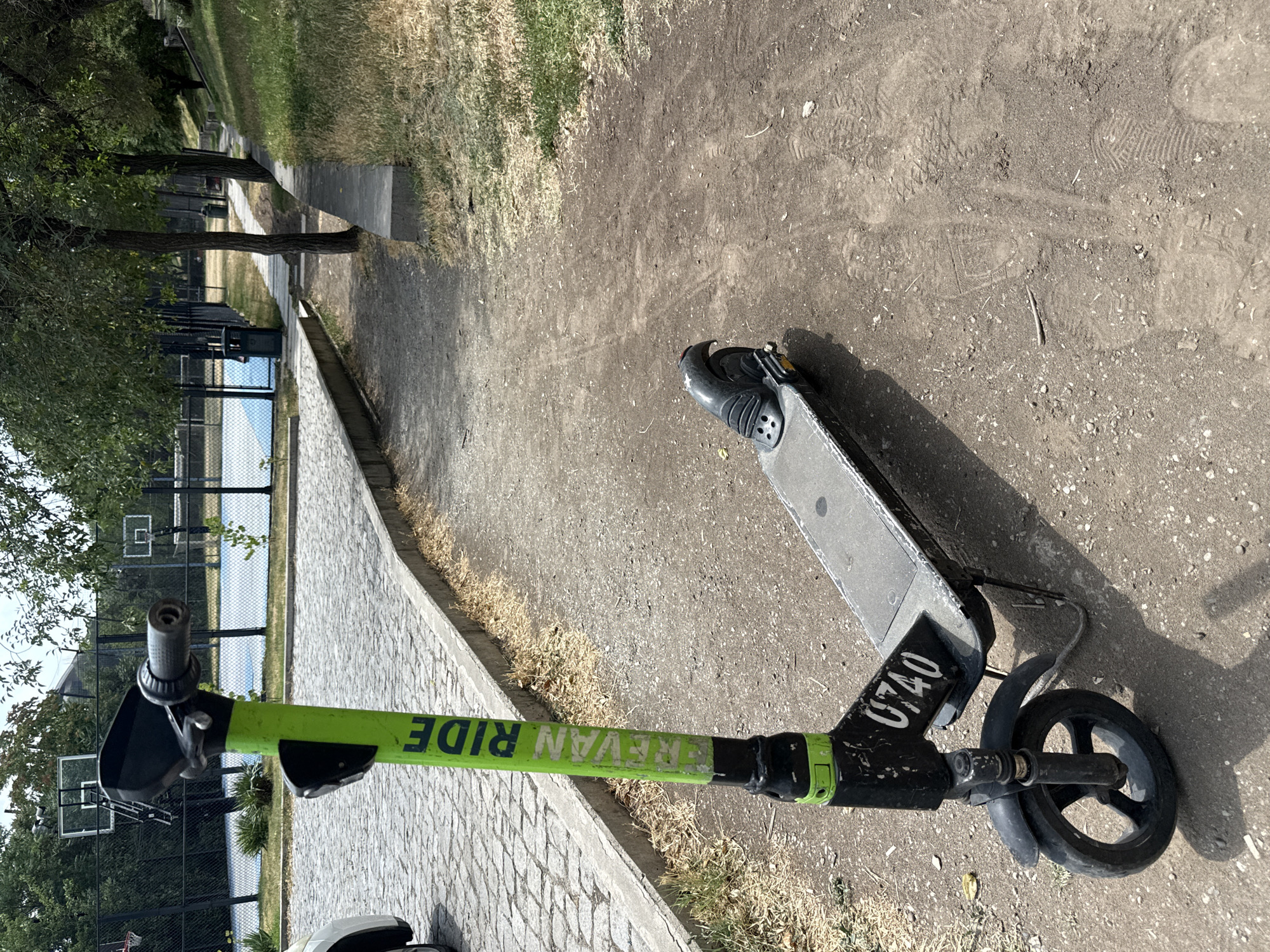 A bright green and black electric scooter with the markings "REVAN RIDE" and the number "0740" is parked on a dirt path next to a cobblestone sidewalk. The scooter is the primary subject of the image, positioned in the foreground and angled slightly to the right. The background depicts a park-like setting with trees, grass, and a basketball court visible beyond a fence. The basketball court appears to be in use, with a blue playing surface and basketball hoops. The environment suggests an outdoor recreational area within a city. The lighting indicates a daytime scene, with distinct shadows cast by the scooter and surrounding objects. The overall atmosphere is one of urban leisure.