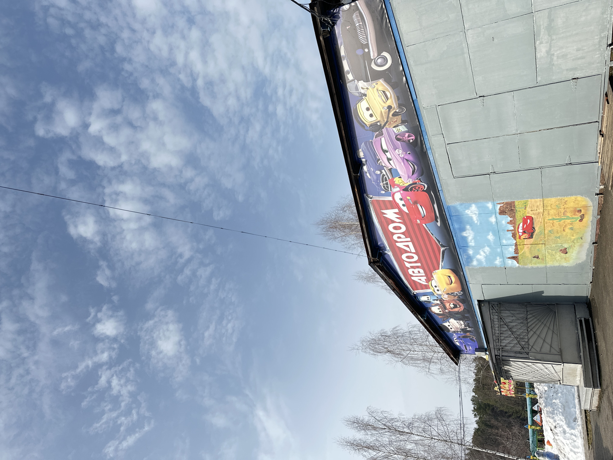 The image depicts a single-story building, likely an amusement ride facility or children's entertainment center, under a clear blue sky with scattered white clouds. The building's exterior is constructed from light grey-green panels, showing visible seams and hinges. Along the upper facade, below the dark-colored roofline, is a large banner featuring cartoon cars from the Disney-Pixar "Cars" franchise. Prominently displayed in red Cyrillic letters on a striped background is the word "АВТОДРОМ" (AVTODROM), meaning "Autodrome" or "Car Track." Characters visible on the banner include Lightning McQueen, Sally Carrera, Mater, Luigi, and a black vintage car. Below the banner, a mural is painted on the building's lower section, depicting a desert landscape with a red car (resembling Lightning McQueen) and cacti.
An entrance on the left side of the building features a double-leaf grey metal gate with a radial, sunburst-like design, accessed by a set of concrete steps. To the left of the entrance, and extending behind the building, are patches of dirty, melting snow on the ground. Bare deciduous trees, appearing to be birch, stand in clusters around the building and in the mid-ground. A dense, dark forest is visible in the distant background. A single dark utility cable or wire crosses the upper right portion of the sky. The ground in the immediate foreground is dark asphalt or dirt. There are no individuals visible in the scene.
This scene is located in Chaikovsky, Russia, likely within a public recreational area such as the city's Park of Culture and Rest, given the nature of the "Autodrome" attraction and the surrounding park-like environment.