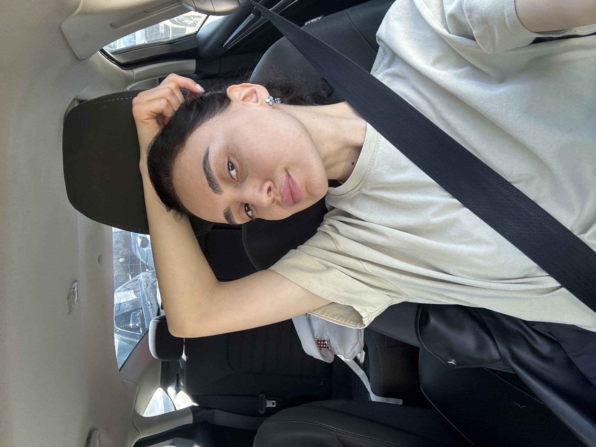A young woman with dark hair pulled back from her face is seated in a car wearing a seatbelt. She is wearing a light-colored t-shirt and black pants. Her right arm is raised, with her hand touching her head. The car's interior is visible, including the seats and a sunroof. The location appears to be in the city of Tbilisi, Georgia, based on exterior clues.