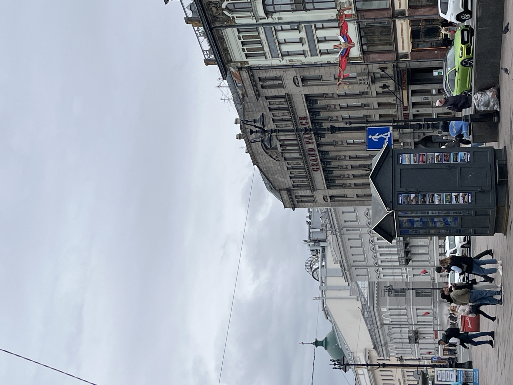 A daytime, eye-level shot captures a vibrant city street under an overcast sky. To the right, a majestic multi-story building stands prominently, featuring classical architecture with numerous windows, columns, and intricate decorative details. The words "GRAND PALACE" are clearly visible on its facade. A flag with white, blue, and red stripes hangs from this building.
In the mid-ground, a dark grey kiosk or newsstand is positioned on the pavement, displaying various images or postcards on its sides. Adjacent to it, a blue pedestrian sign depicting a walking figure is mounted on a pole. A bright green car is parked on the street, with a white car visible behind it. A person in a dark jacket and a light-colored hat stands near the green car, next to a trash bag.
Numerous pedestrians populate the scene. A group of casually dressed individuals walks from left to right further down the street, past the kiosk. Another smaller group is visible in the distance. On the far left, the dome or spire of another building, possibly green, is partially visible. Overhead power lines or tram wires stretch across the uniform grey sky, adding to the urban feel. The scene depicts a bustling city environment, likely in a European setting, under calm, cloudy weather.