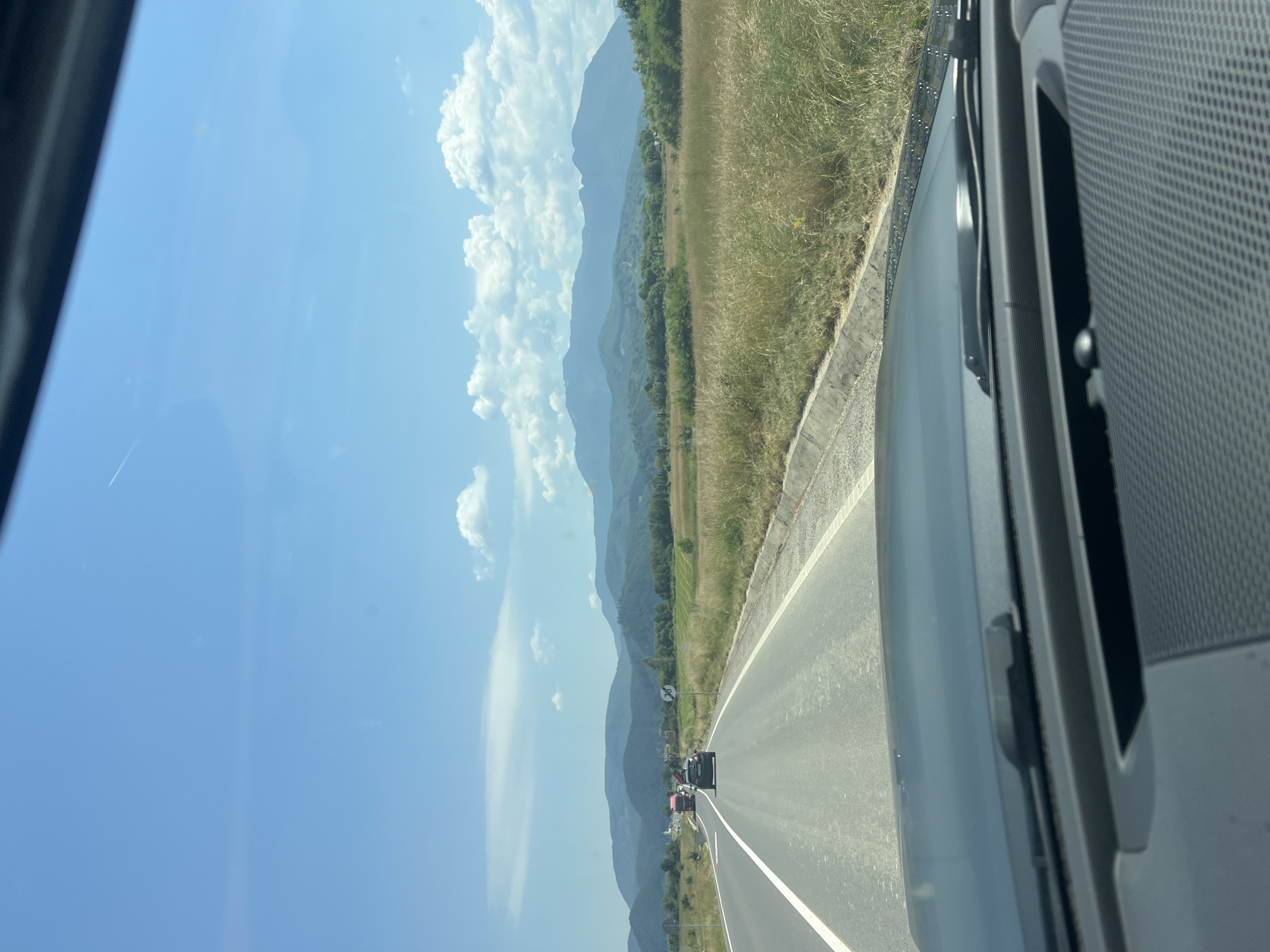 A vertical, eye-level shot shows a view from the interior of a vehicle traveling on a highway. The road is paved and marked with white lines. To the right of the road, a dry, grassy field stretches into the distance. Beyond the field, a line of trees forms a green border, behind which rolling, hazy blue mountains rise against a clear blue sky dotted with fluffy white clouds. The sun is bright, indicating it is daytime, likely mid-morning or afternoon given the sunlight.
In the distance on the highway, several vehicles are visible, including a black SUV and some red trucks, suggesting moderate traffic. The perspective is from the passenger seat, looking out the window. Part of the vehicle's interior is visible in the foreground on the right side, including a dark gray, textured surface and a portion of a window frame. There is no visible text in the image. The overall mood is serene, capturing a typical travel scene on a bright, clear day with scenic mountainous surroundings.