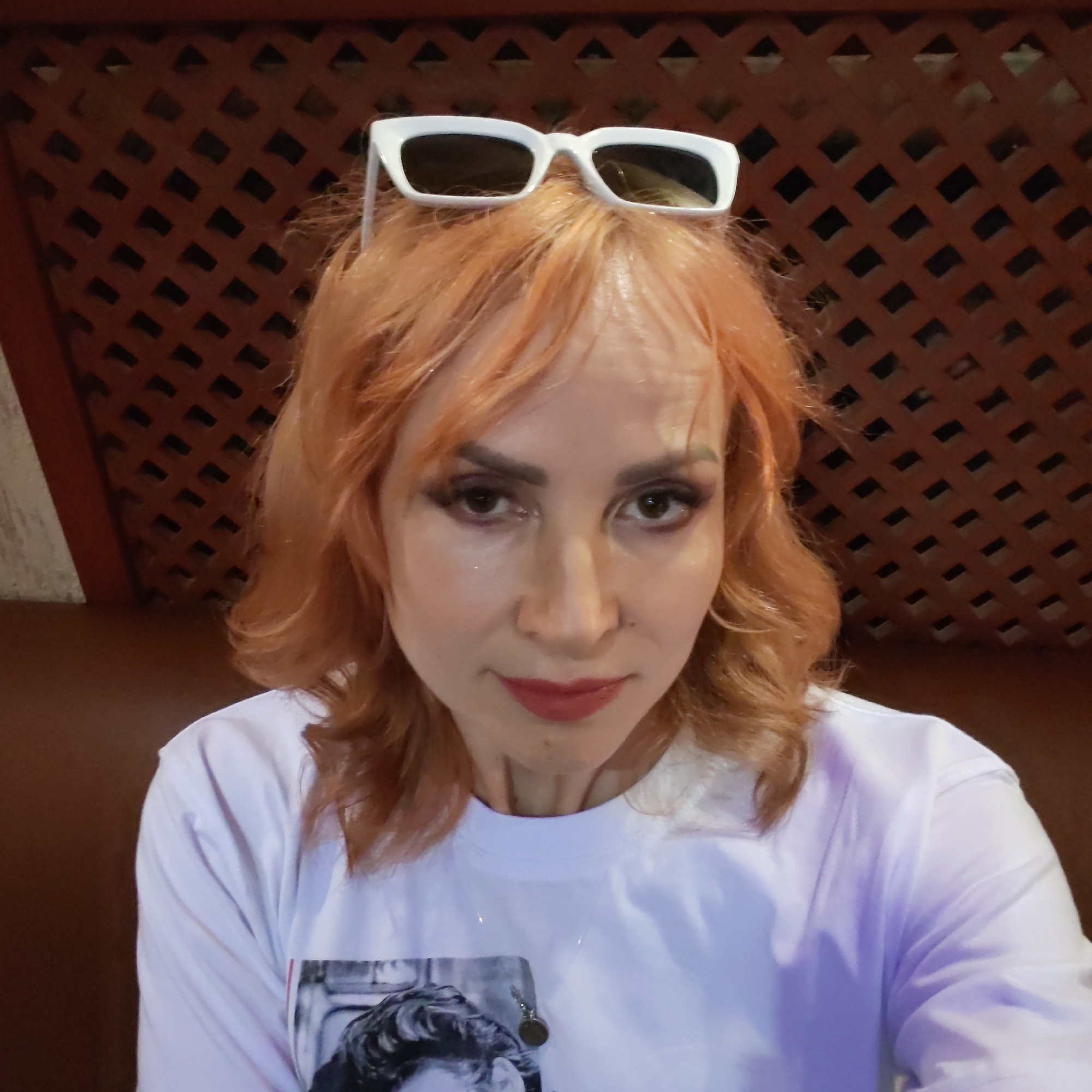 This detailed image captures a woman indoors in Chaikovsky, Russia. The subject, an adult female with medium-length, wavy, reddish-orange hair, is depicted from the chest up, looking directly into the camera. White, rectangular-framed sunglasses are positioned on the crown of her head. Her face exhibits applied cosmetics, including prominent dark eye makeup, defined eyebrows, and red lipstick; a small dark mark is visible near the right side of her nose. She is wearing a white crew-neck t-shirt that displays a partially visible black-and-white graphic print of an individual's face on the front, and a thin necklace with a small, dark, circular pendant around her neck. The background is characterized by a dark reddish-brown wooden lattice panel arranged in a diamond pattern, situated above a solid brown surface. Artificial illumination produces highlights on the subject's skin and a subtle purple coloration on the right sleeve of her shirt.