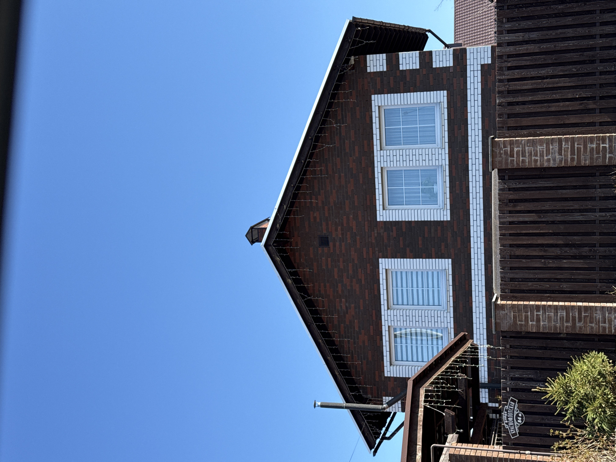 The image depicts the exterior of a brick house under a clear blue sky. The house is primarily built with dark brown bricks contrasted by white brick outlines around the windows. The house has a gabled roof and two stories visible, with three windows. A wooden fence or structure is visible to the right of the house. A sign labeled "ул. КРАСНОФЛОТСКАЯ, 84а" (Krasnoflotskaya Street, 84a) is seen attached to the fence. Based on the sign, the location is inferred to be 84a Krasnoflotskaya Street in Votkinsk, Russia. There are no people visible in the image. The house is decorated with lights hanging from the eaves.