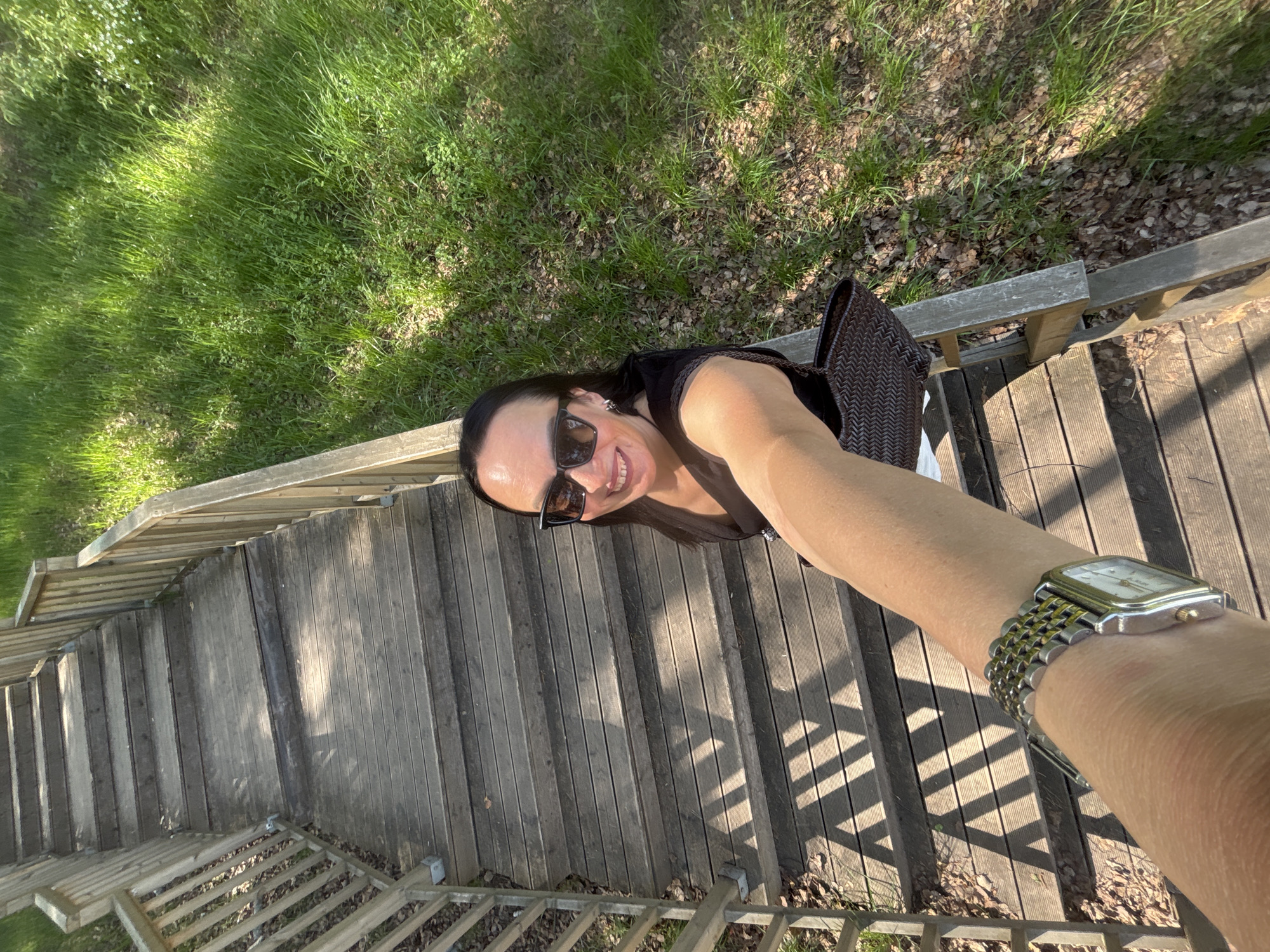 A woman stands on wooden stairs in Chaikovsky, Russia, taking a selfie. She is wearing sunglasses and a dark top. A brown woven bag is slung over her shoulder. Her arm is extended, showing a silver and gold watch. The stairs have wooden railings and lead down to a grassy area with some dead leaves.