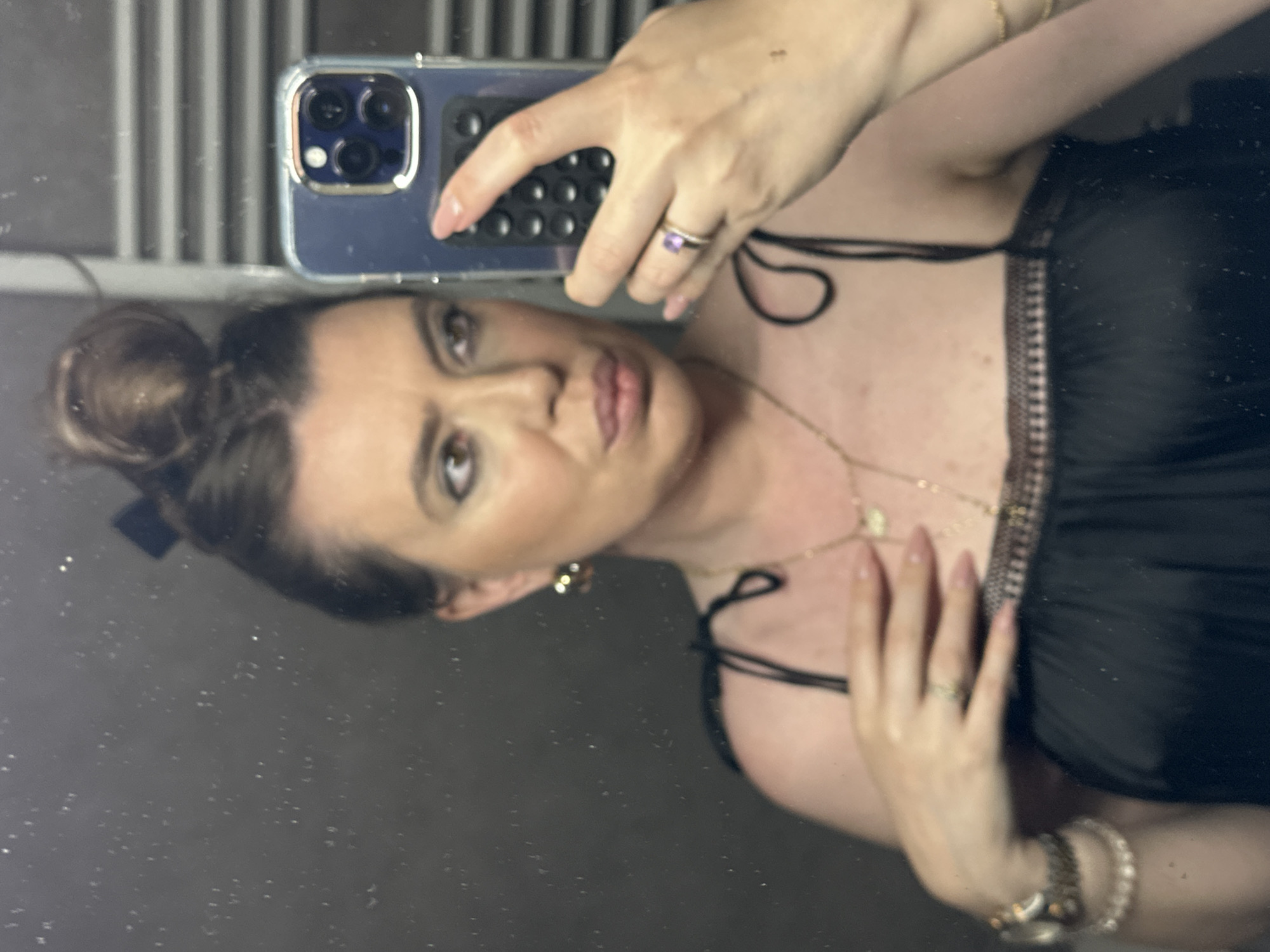 A mirror selfie, rotated 90 degrees clockwise, captures a woman from the chest up. She appears to be an adult with dark hair tied in a bun, adorned with a dark bow. Her face shows makeup, including defined eyebrows, eyeliner, and lipstick, with a relaxed, confident expression. She holds a smartphone, resembling an iPhone with multiple cameras, in a clear case with her right hand. Her left hand rests gently against her chest area.
She is wearing various pieces of jewelry: a ring with a purple gemstone on her right index finger, a delicate gold-toned necklace with a small pendant, matching earrings, and a watch layered with bracelets on her left wrist. Her attire consists of a black top or dress with thin straps and a textured or ruffled trim across the chest. The reflection shows vertical stripes in the background, possibly from blinds or a wall, and the mirror surface itself appears slightly speckled. The activity is clearly a self-portrait taken in a mirror. No specific time of day or weather is discernible, and there are no visible location cues within the image itself to confirm the provided context of Tbilisi, Georgia. No legible text is visible.